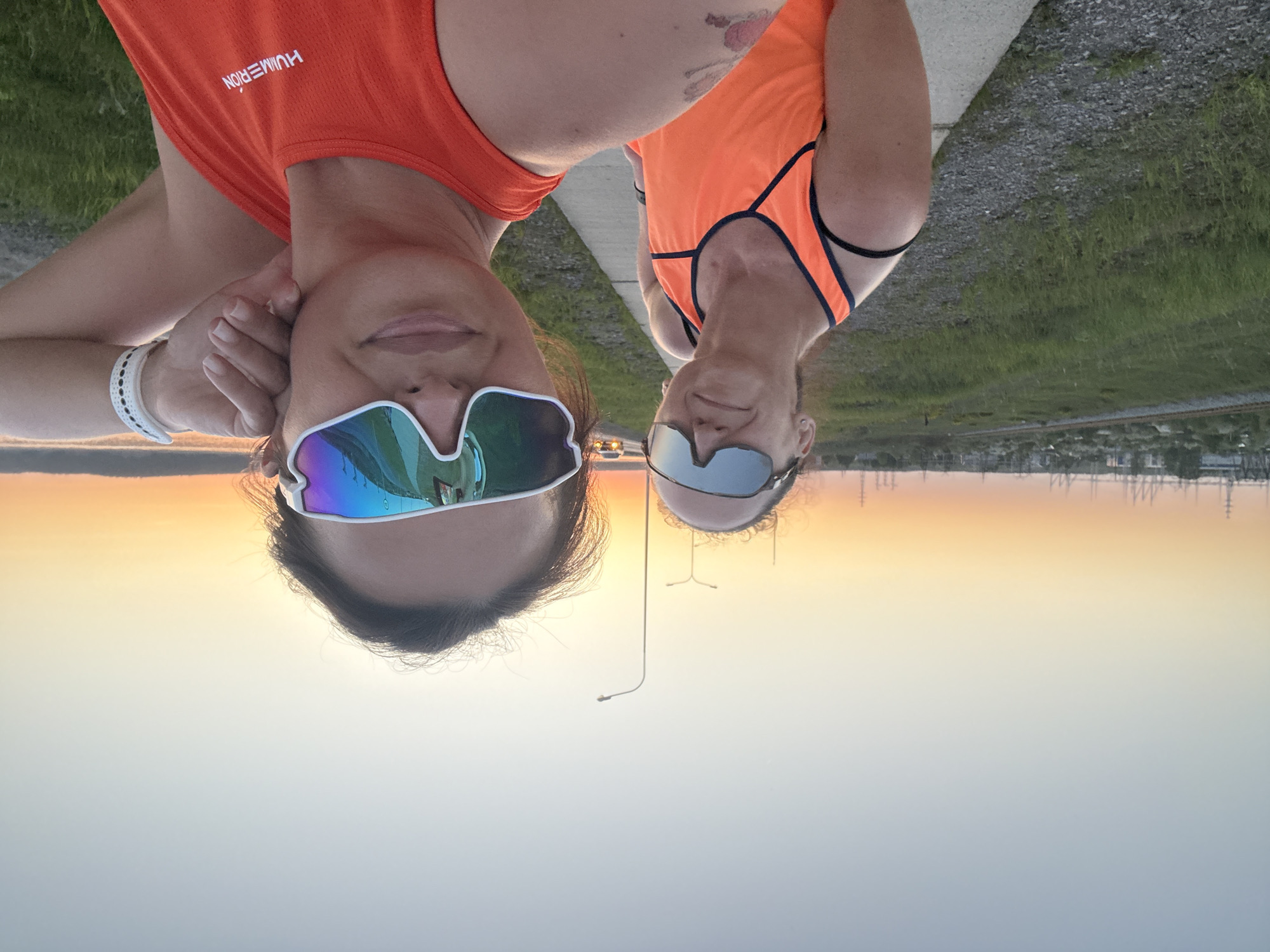 Two women in athletic wear pose for a selfie outdoors at sunset in Chaikovsky, Russia. The woman on the left wears a bright orange tank top with white lettering, white sunglasses, and a white wristband. The woman on the right wears an orange tank top with black trim and dark sunglasses. The backdrop features a concrete path with grass on either side, a distant cityscape silhouette against the sunset sky, and power lines.