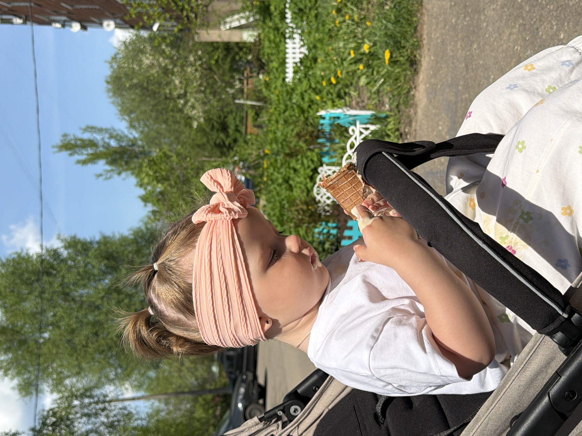 A young child sits in a stroller, eating an ice cream cone. The child has fair skin, brown hair pulled back in pigtails, and a pink headband. The child is wearing a white shirt. The stroller is black and gray, with a white patterned blanket. The background includes trees, a blue and white fence, and a building. The image was taken in Reshetnikovo, Russia.