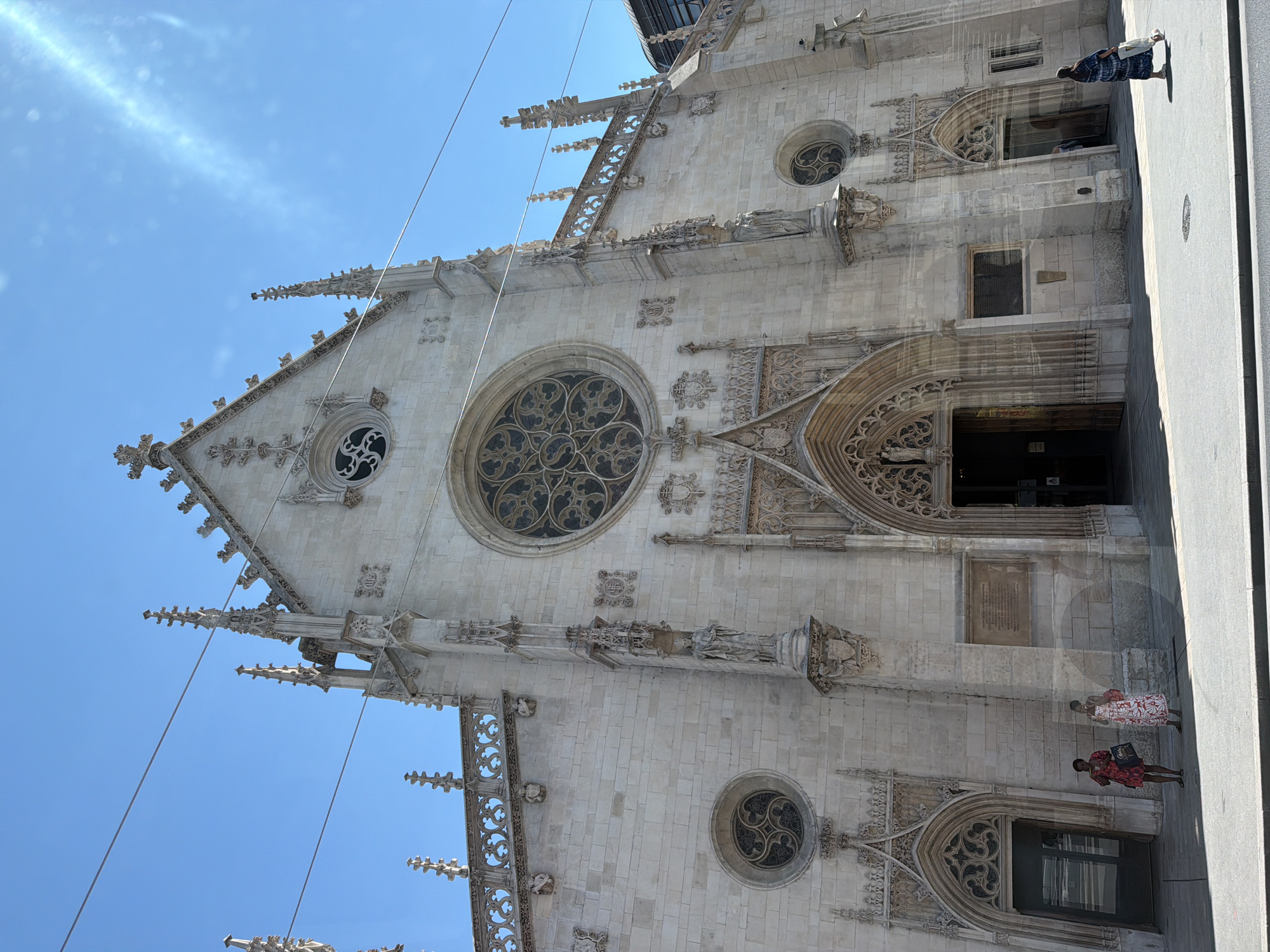 This media file is a vertically oriented photograph of the exterior of a large, ornate cathedral, likely in Lyon, France. The building's facade is constructed of light-colored stone with intricate Gothic architectural details, including pointed arches, spires, gargoyles, and a large rose window. A smaller circular window with a similar decorative pattern is visible lower on the facade. Two women are standing on the sidewalk in front of the cathedral, their backs to the camera. One woman is wearing a patterned dress and carrying a bag, while the other is wearing a red dress. A third person, dressed in dark clothing, is walking away from the camera further up the street. The sky is bright blue with some wispy clouds, suggesting it is daytime and the weather is clear. Several thin wires crisscross the upper portion of the image, possibly power lines or utility cables. The overall impression is of a sunny day in a city with historical architecture.
