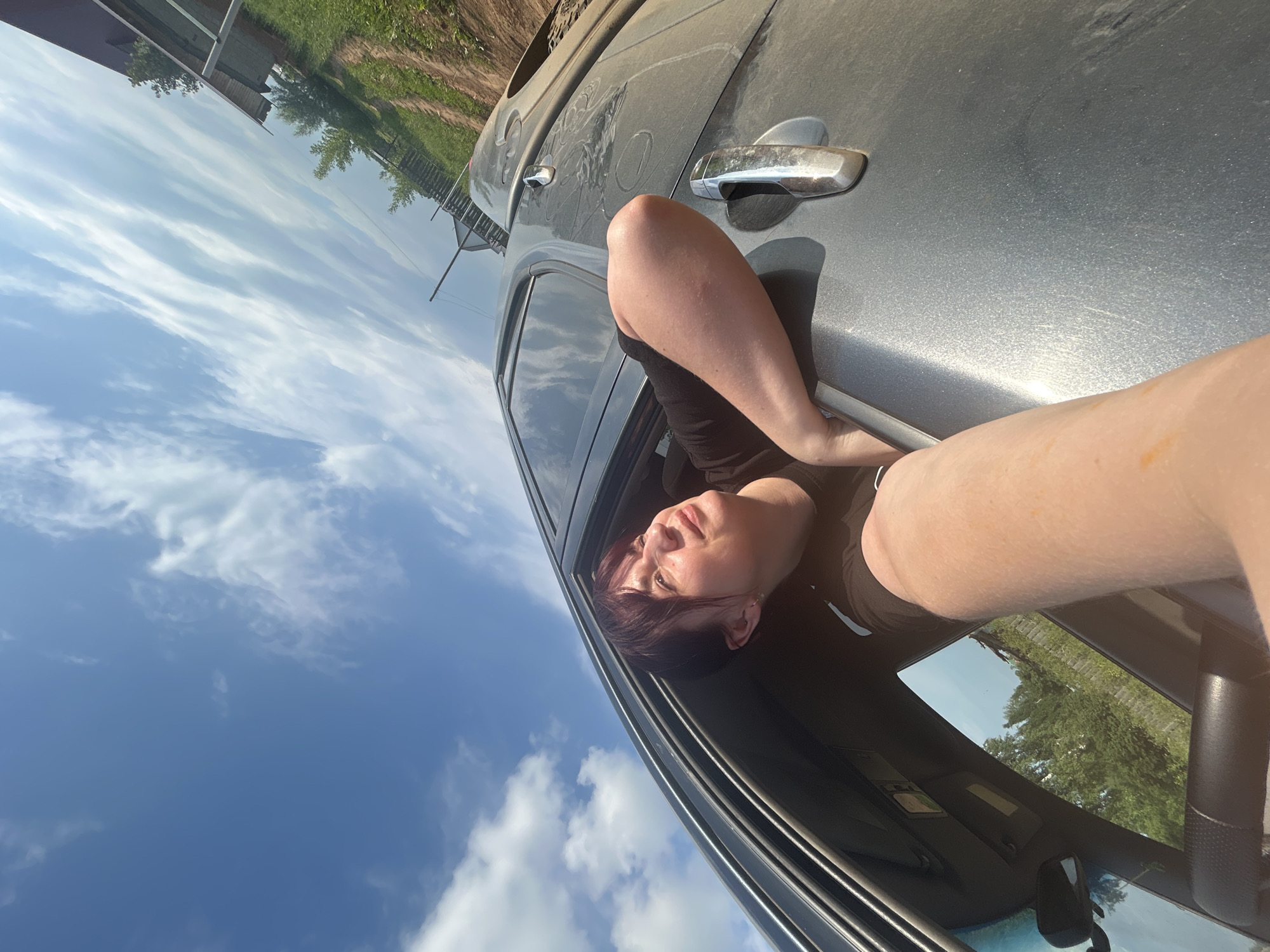 A person is leaning out of a car window, looking up at the sky. The car appears to be on an unpaved road in a rural area. The person has dark hair and is wearing a black t-shirt. The sky is blue with some white clouds. Trees and a fence can be seen in the background. The overall impression is a snapshot of a person enjoying a moment outdoors.