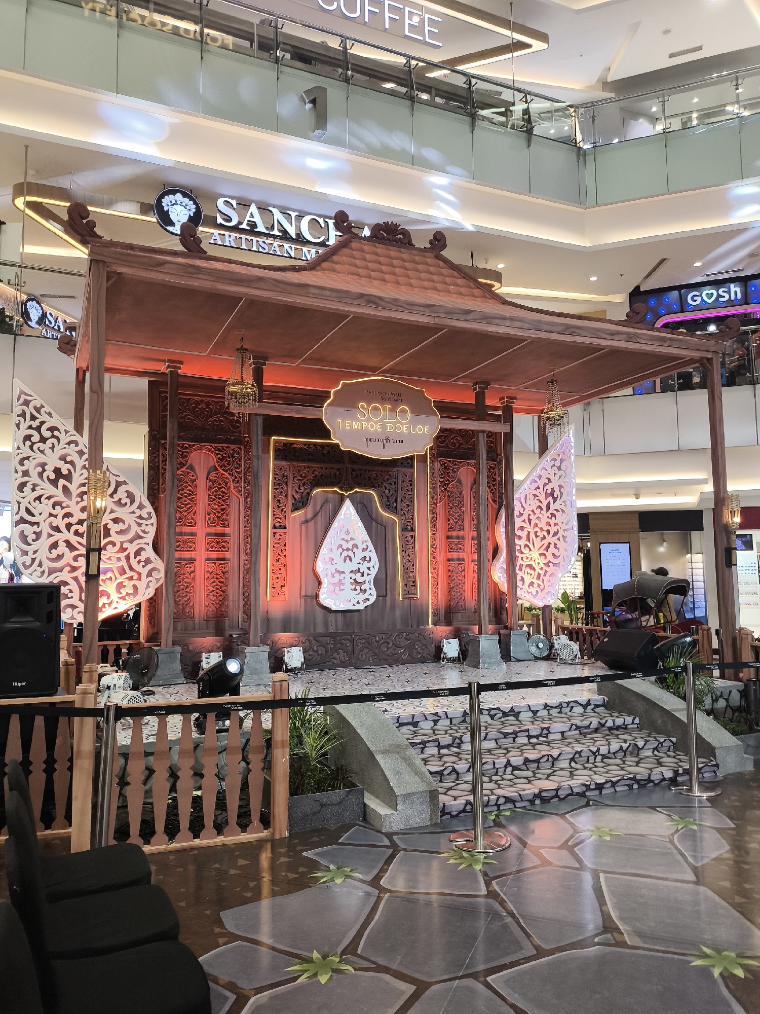 In the city of Grogol, Indonesia, an indoor stage is set up within a shopping mall, possibly for an event or exhibition. The stage is adorned with traditional Indonesian architectural elements, featuring intricately carved wooden panels and a tiered roof structure. Two large, white, ornate panels resembling Wayang Kulit figures are positioned on either side of the stage. The backdrop of the stage is a detailed wooden carving, illuminated by red lighting. Above the stage, a sign reads "SOLO TEMPOE DOELOE" in a decorative font, suggesting a theme related to the past or heritage of Solo. Chandeliers and various lighting equipment, including spotlights and fans, are visible on and around the stage, indicating preparations for a performance or display. Speakers are placed on the left side of the stage, and a small rickshaw-like vehicle is parked to the right. The floor in front of the stage is decorated with large, grey, stone-like tiles and green leaf motifs. A barrier, consisting of wooden railings and stanchions with black ropes, separates the stage area from the viewing area, where several empty black chairs are neatly arranged. In the background, shopping mall signage, including "SANCH ARTISAN M" and "gosh," can be seen, confirming the location as a commercial space. The overall scene conveys a cultural theme presented in a modern, public setting.