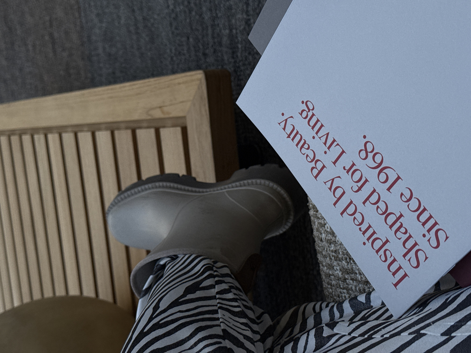 The image shows a person sitting in a chair with a white boot and zebra-striped pants. Next to the chair is a book with the text "Inspired by Beauty. Shaped for Living. Since 1968." written in red. The location is not specified in the image.