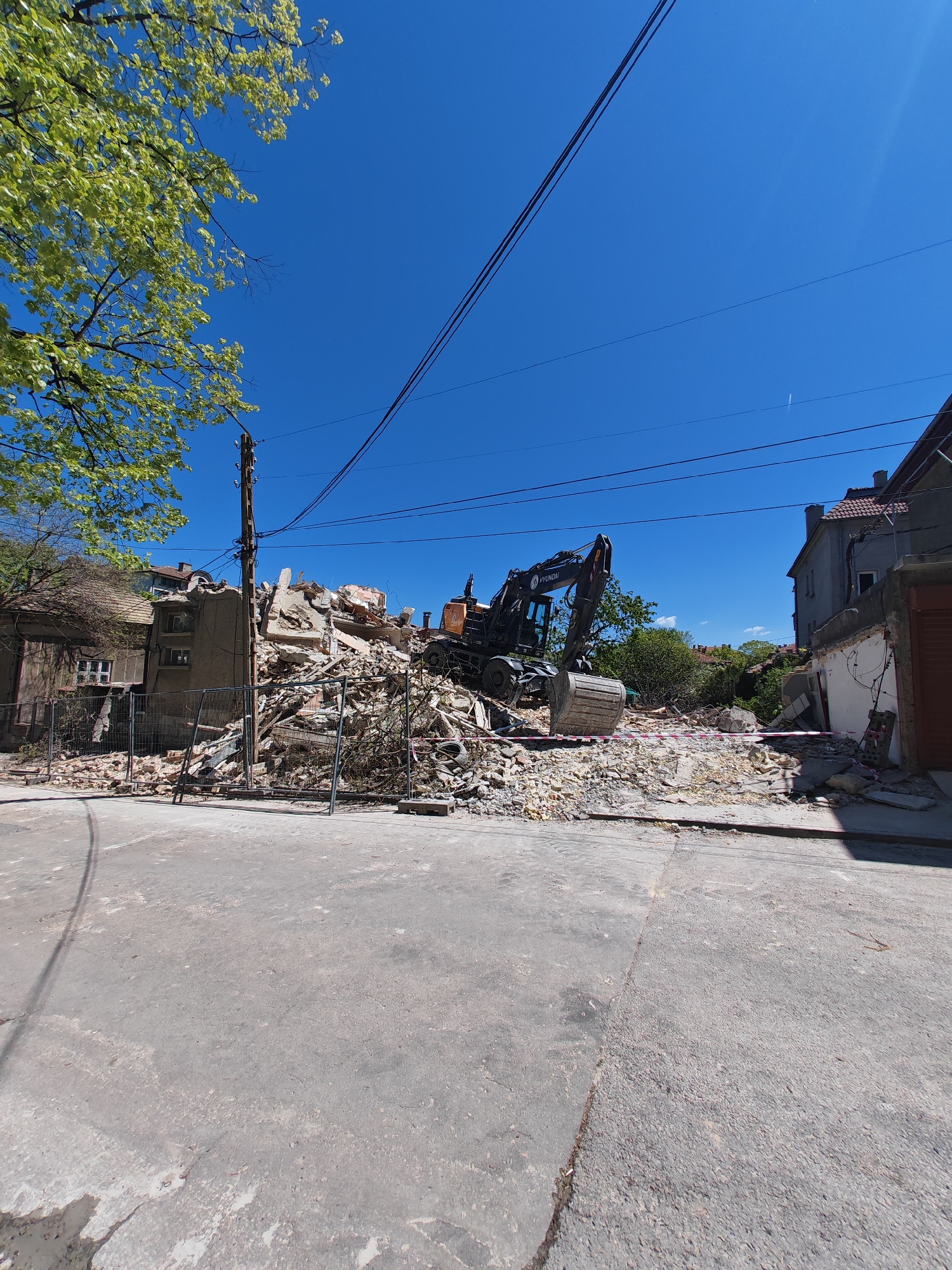 The image shows a demolition site in what appears to be a residential area in Shumen, Bulgaria. A large pile of rubble fills the space where a building once stood, partially enclosed by a temporary fence. A dark-colored Hyundai excavator is positioned atop the debris. Adjacent buildings remain standing, with one partially visible to the left and another to the right. The street in the foreground is paved and grey. Power lines run overhead against a clear blue sky. A tree with green foliage is visible on the left. Pink and white barrier tape lies on top of the rubble.