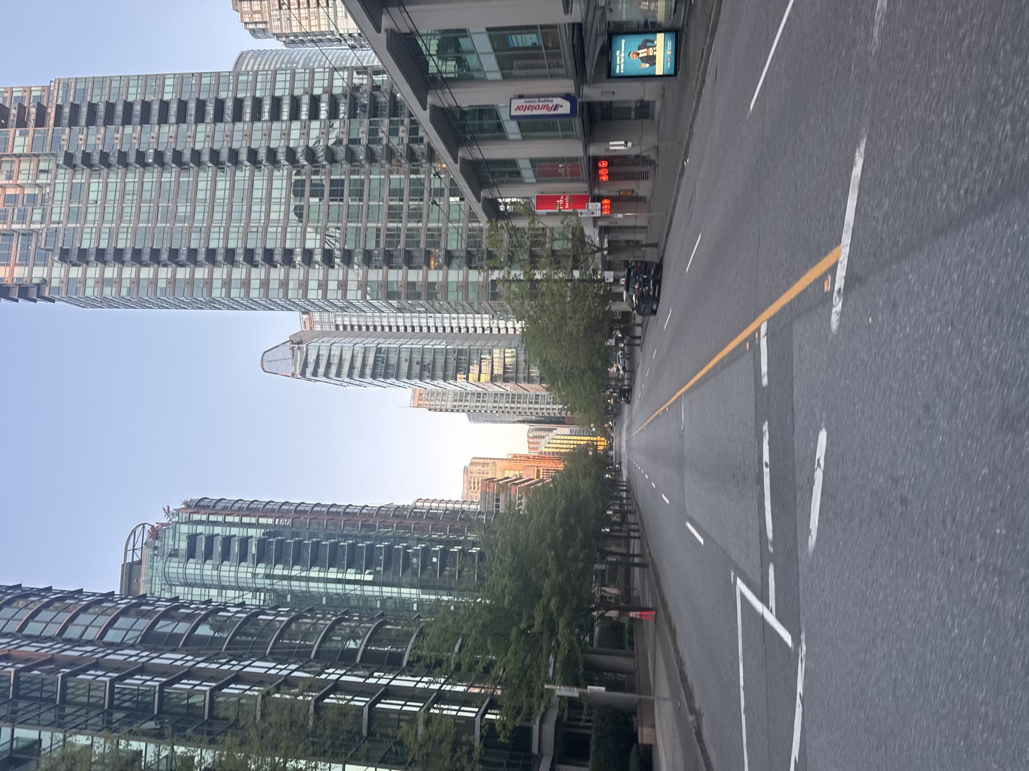 The image depicts a straight, wide city street lined with modern, high-rise buildings, most of which appear to be residential. The buildings feature reflective glass facades and stepped designs. A row of trees flanks the sidewalk along the right side of the street. The street is marked with white and yellow lane markings. Several vehicles are visible in the distance. Signage for "Purolator" and a sushi restaurant are present on one of the buildings. The scene suggests downtown Vancouver, Canada.