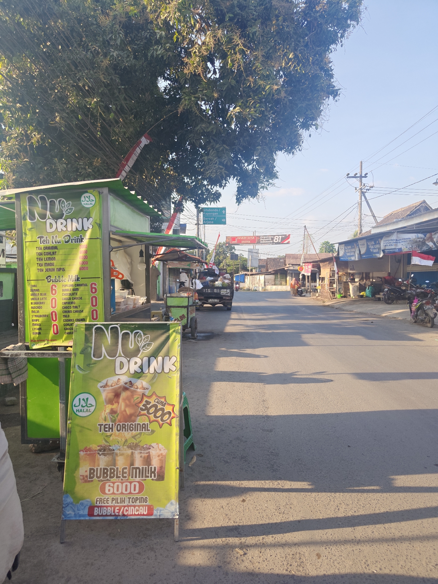 A street scene in Klampokan, Indonesia, captures a vibrant commercial area under a clear blue sky. A Nu Drink stall, identifiable by its green awning and prominent signage, is set up on the left side of the road, offering a menu of teas and bubble milk drinks. The menu lists various flavors like Teh Original, Teh Coklat, and Teh Lemon, with prices indicated. A second, smaller Nu Drink sign stands in front of the stall.
Across the street, a row of small businesses lines the road, with motorcycles parked in front. A black pickup truck is visible further down the street, and pedestrians can be seen walking. Various banners and flags, including Indonesian national flags, are displayed on poles and buildings, suggesting a festive atmosphere or ongoing celebration. Overhead, power lines crisscross the sky, typical of an urban or semi-urban setting. The road surface appears to be asphalt, with shadows cast by trees and structures indicating the time of day. The overall impression is one of a lively local market or street with active commerce.