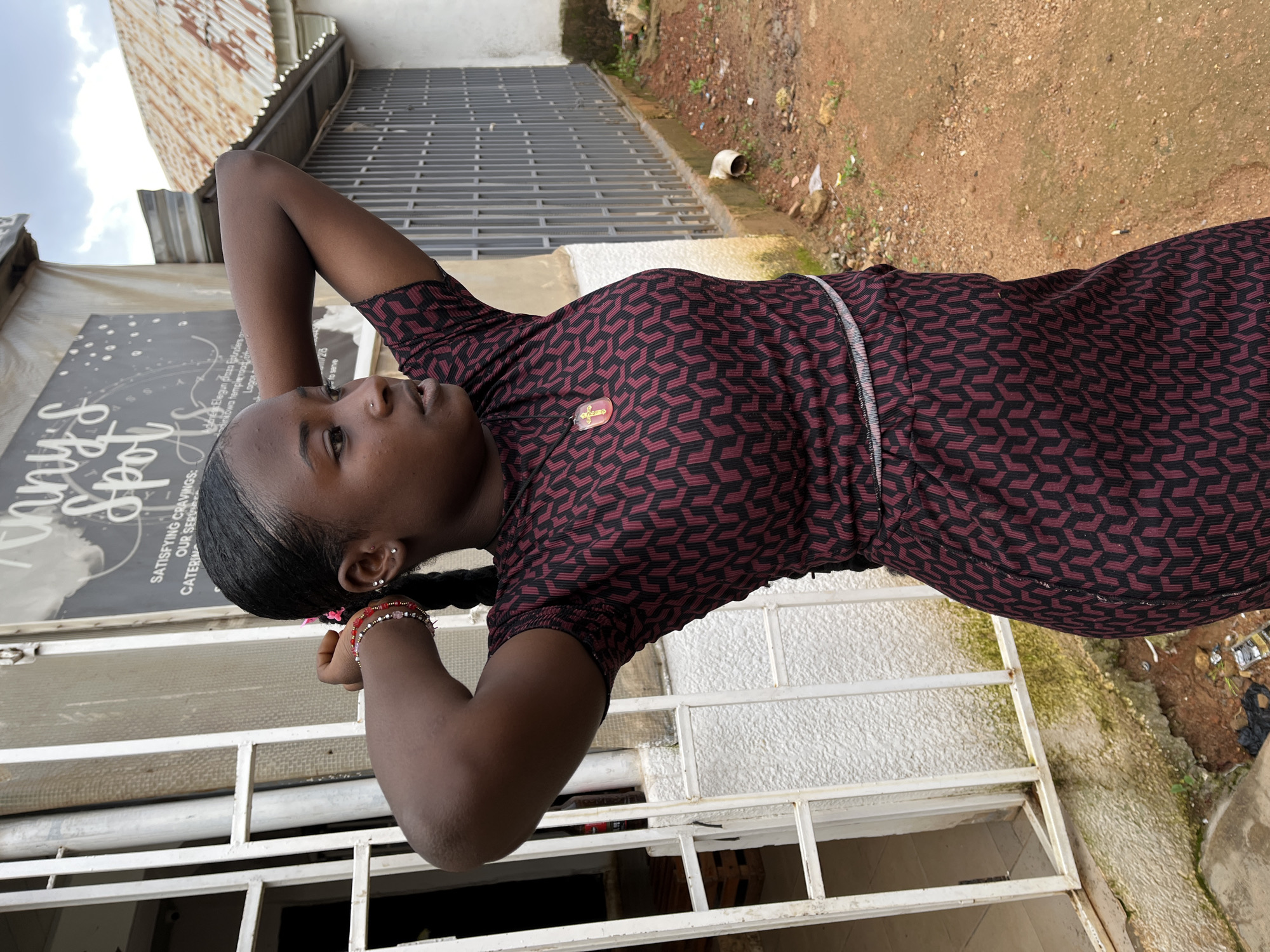 A young Black girl with dark hair braided down her back is standing with her arms raised behind her head. She is wearing a maroon and black patterned dress with a thin silver belt. On her dress, just above her waist, is a small gold pendant shaped like a cross with a brown stone in the center. She has two earrings in her left ear and a red and pink beaded bracelet on her left wrist. Her expression appears thoughtful as she looks to the left, away from the camera.
Behind her, a metal grate covers a window or opening. To the left of the girl, a black sign with white lettering is visible. Text on the sign reads: "Sunny Spot", "SATISFYING CRAVINGS:", and "OUR SERVICES: CATERING, RES...". To the right of the girl, there is a dirt path with some rocks and sparse green vegetation. A white pipe protrudes from the ground.
The scene appears to be outdoors, possibly in a semi-urban or residential area, given the building materials and the surrounding environment. The lighting suggests it is daytime, with overcast skies. The overall mood is casual and relaxed.