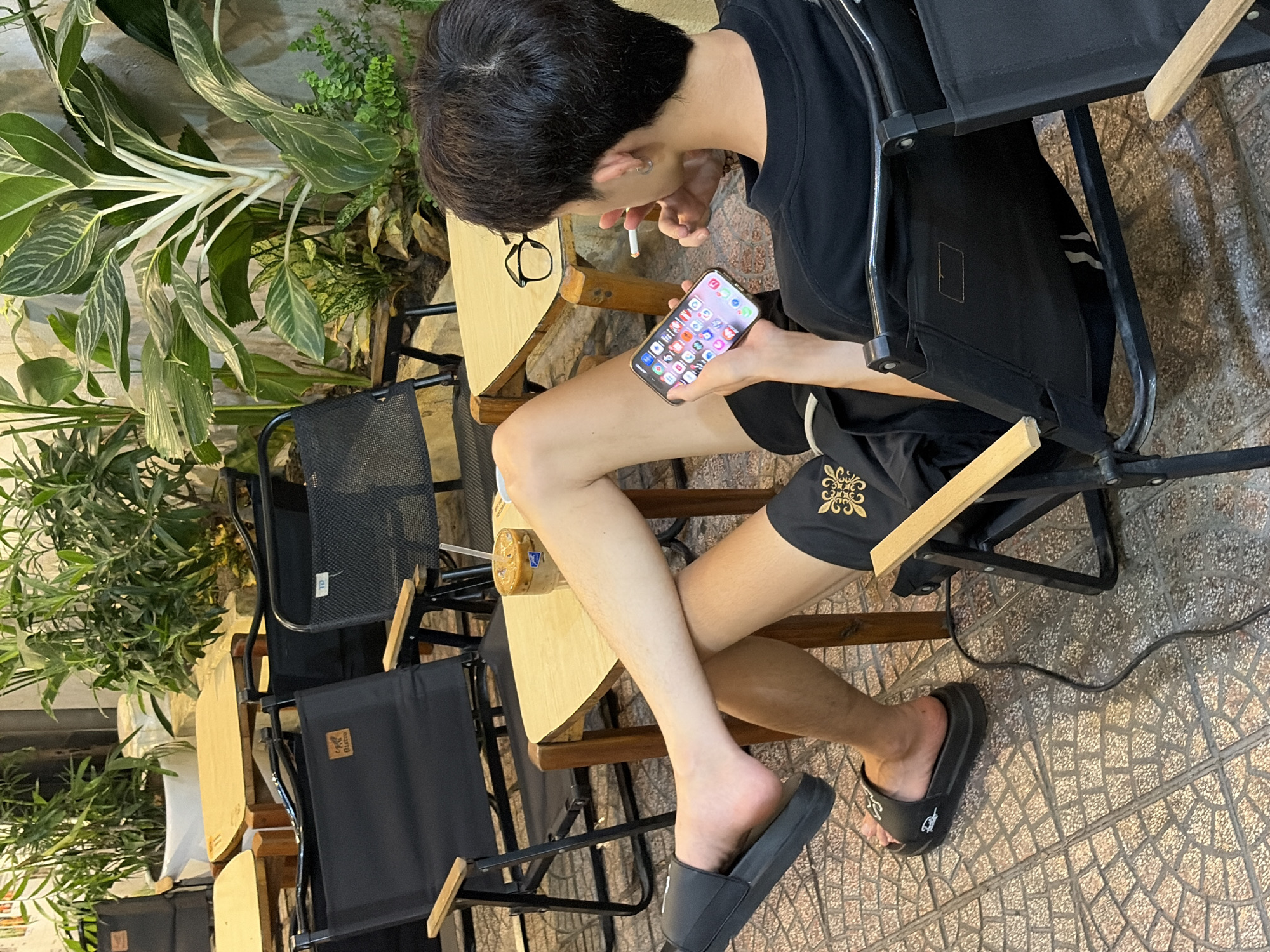 A young East Asian male sits in a black folding chair at an outdoor cafe in Da Nang, Vietnam. He is wearing a black t-shirt and black shorts, and black slides on his feet. He holds a smartphone in his left hand, and a cigarette in his right hand, which is near his mouth. A pair of glasses rests on a light-colored wooden table next to him. A glass of iced coffee with a straw sits on the table in front of him. The background shows several potted plants and more black folding chairs and tables. The ground is paved with patterned tiles.