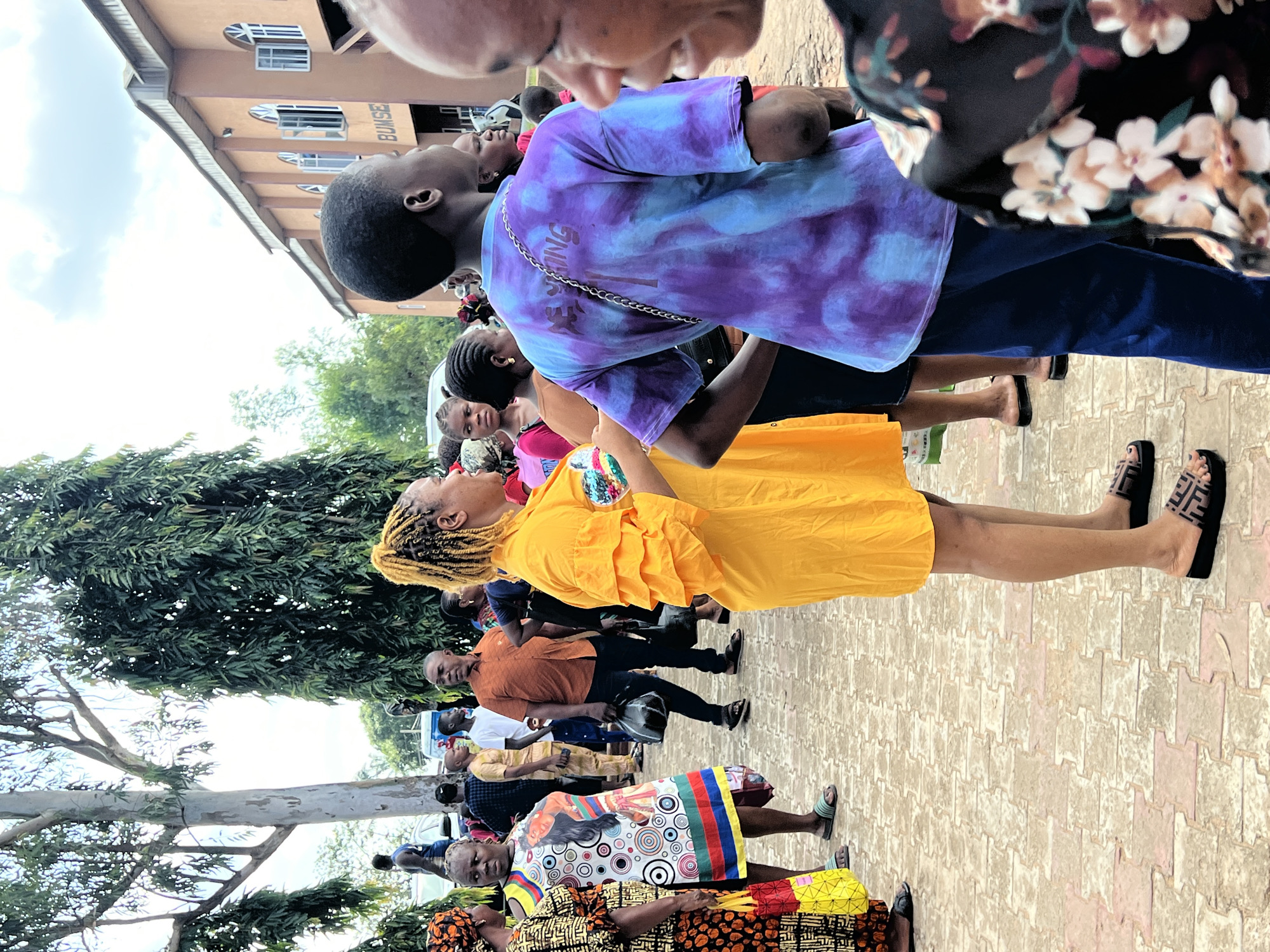 The image shows a group of people standing outside a building with "BUMSE" written above the entrance. The building appears to be made of tan brick with arched windows. In the foreground, a woman in a bright yellow dress stands facing right, her hair in a blonde ponytail. To her left, another woman wears a colorful, patterned dress. Further back, a man in an orange shirt is visible. The ground is paved with interlocking bricks. Tall trees with dense green foliage are present on the left side of the image, with a lighter-colored, thinner tree trunk also visible. The sky is partly cloudy. The scene appears to be an outdoor gathering or event in Nnewi, Nigeria.
