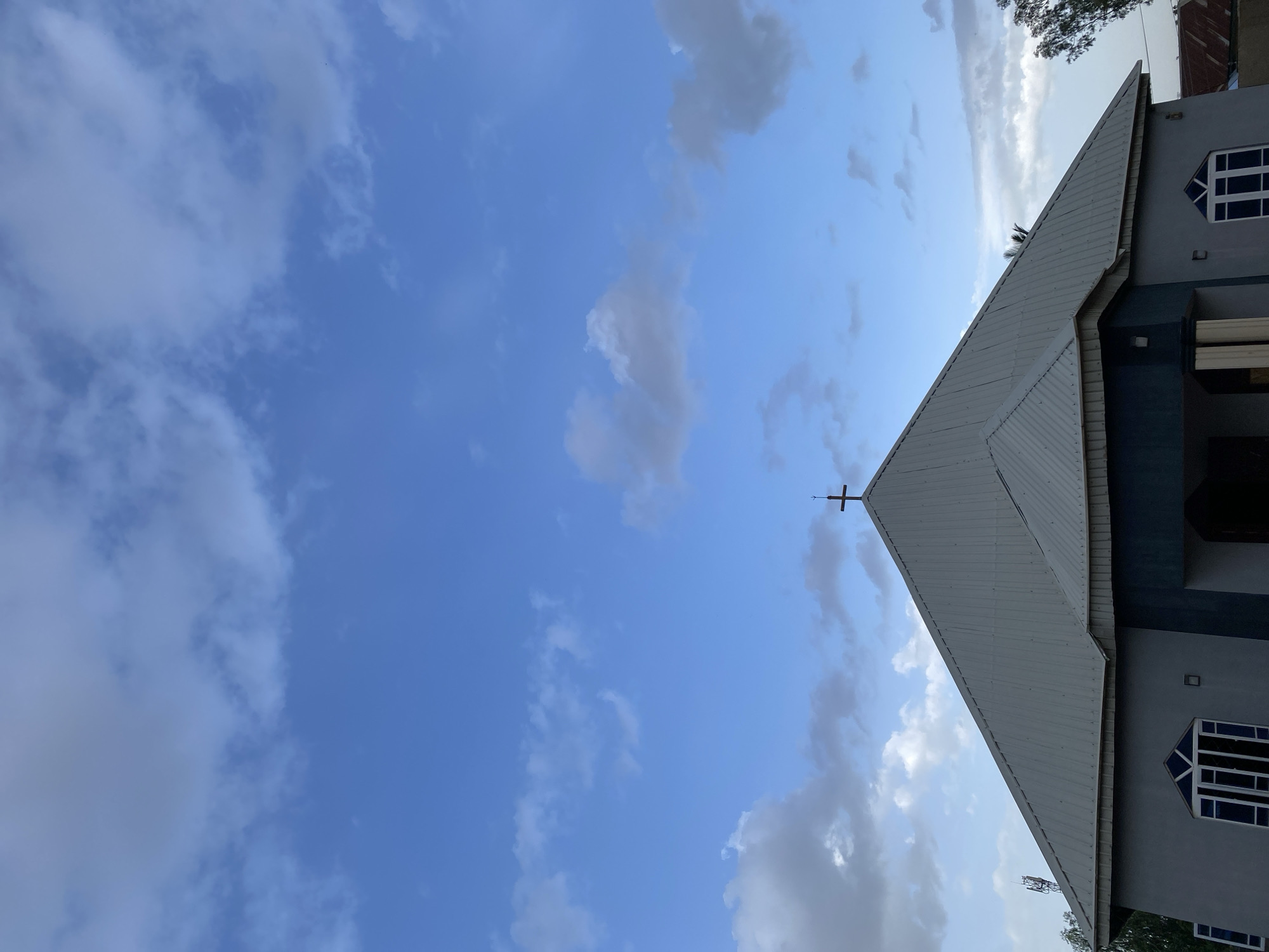 In Jos, Nigeria, the image captures the upper facade of a gray church building against a blue sky dotted with white clouds. A prominent metal-clad, steeply pitched roof dominates the top portion of the structure, culminating in a cross at its apex. The building features several windows with white frames and a dark blue recessed entrance area. To the left of the church, a cell tower is visible, and to the right, the upper branches of a tree. The overall scene suggests an outdoor setting in a built environment.