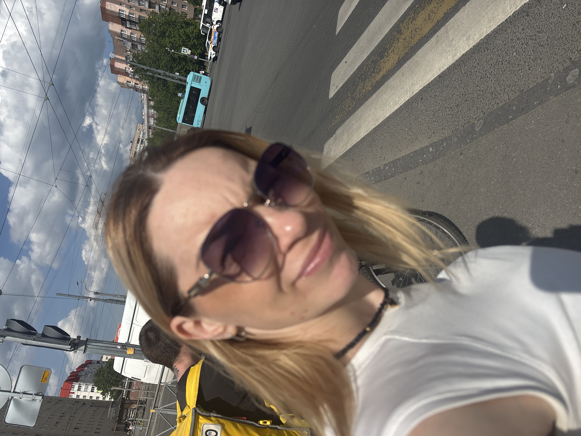 The image captures a first-person perspective, likely a selfie, of a blonde-haired woman wearing sunglasses and a white top, with an urban street scene in Saint Petersburg, Russia, as the backdrop. The woman's face is partially visible and slightly out of focus, showing a hint of a smile. To her left, the back of a man's head and shoulder are visible, and he is wearing a bright yellow backpack or jacket.
The setting is an active city street during the daytime, under a bright, partly cloudy sky. In the middle ground, a prominent zebra crossing spans the asphalt road. Various vehicles are present, including a light blue public bus and a white service vehicle, possibly police, with faint Cyrillic text "ПОЛИЦИЯ" (POLICE) and stripes visible on its side. Overhead, numerous power lines, typical for trolleybuses, crisscross the frame. Multi-story buildings with characteristic architecture line the street in the background. Traffic lights are also visible on the left. The overall scene depicts a sunny, casual moment in an urban environment.
