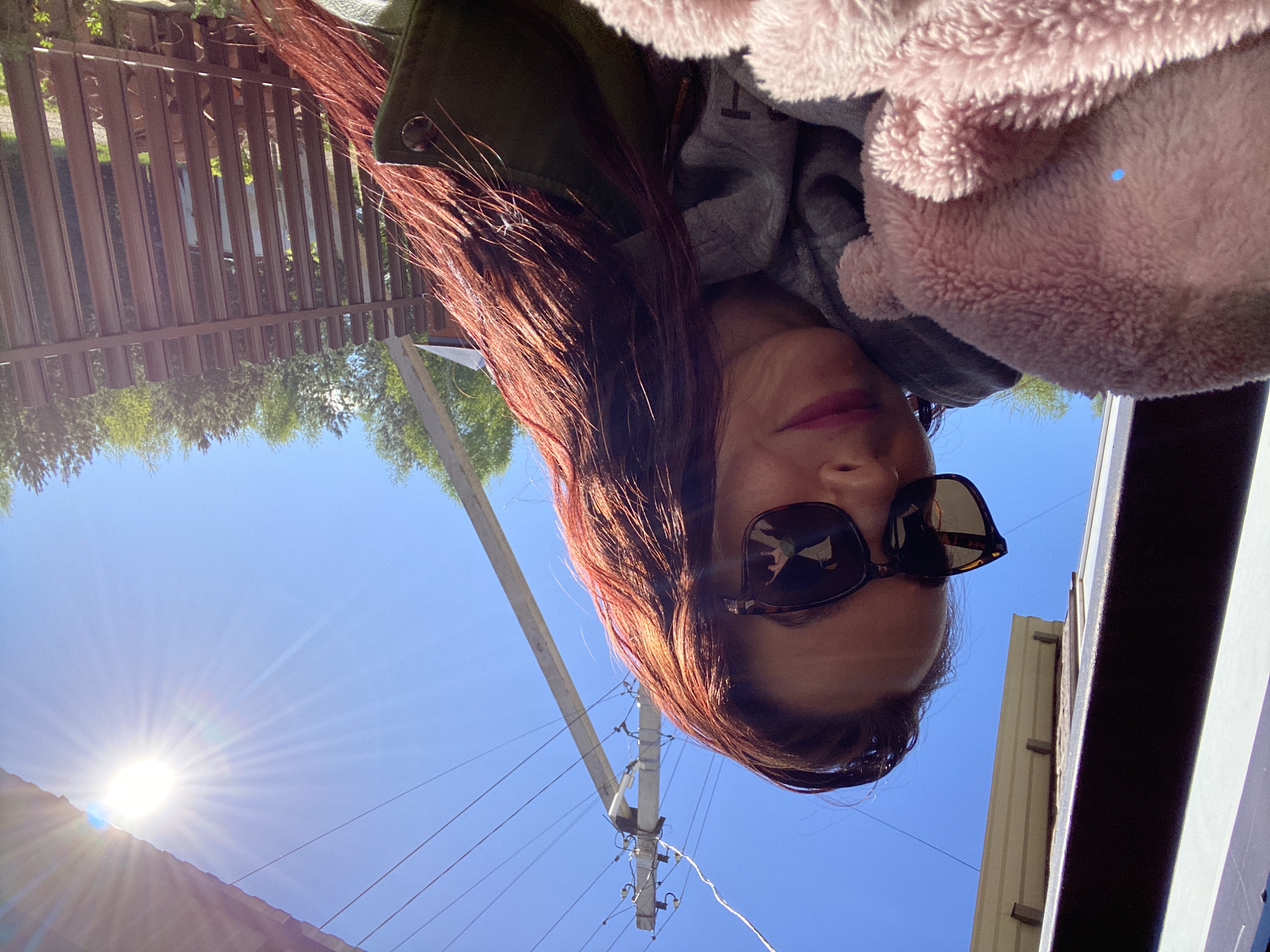 A person wearing sunglasses, a grey hoodie, and a green jacket with a pink fuzzy collar is facing the camera in an outdoor setting under a bright blue sky. The sun is visible in the lower left corner of the image. Behind the person, power lines are strung across an electric pole. A wooden fence is visible in the upper left corner. The location is likely near or in Фоки, Russia, based on the user prompt.