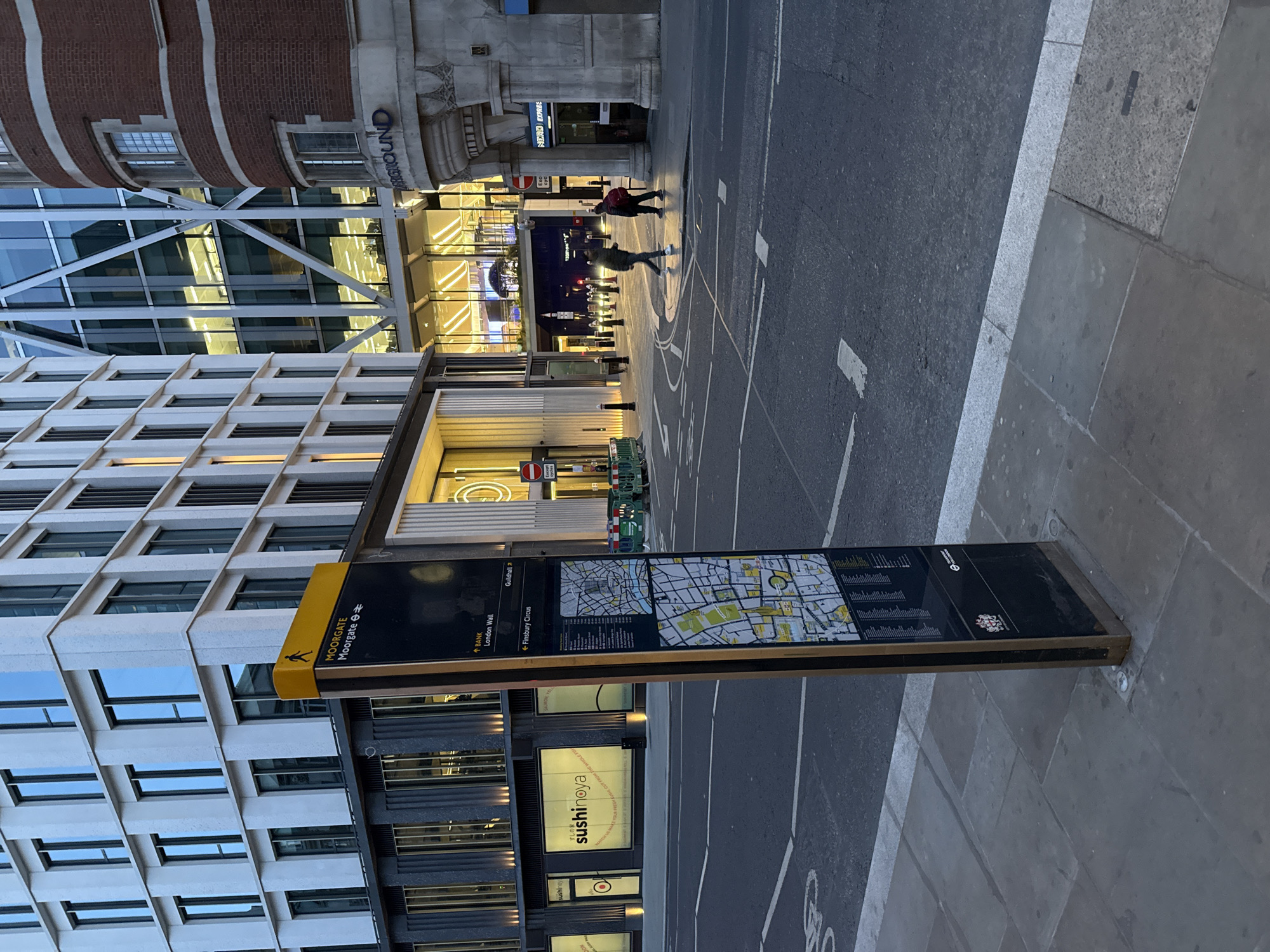 The image captures an urban street scene in London, United Kingdom, featuring a wayfinding sign indicating directions to Moorgate station, Bank, and Finsbury Circus. The sign is located on the pavement alongside a road with pedestrian and cycle markings. Buildings are visible, including a modern office building with multiple windows and a building with "GROUND" in the exterior. Pedestrians are present in the background, walking towards a well-lit area with shops and a "Nero Express" sign. A "sushinoya" restaurant is also visible below the map.