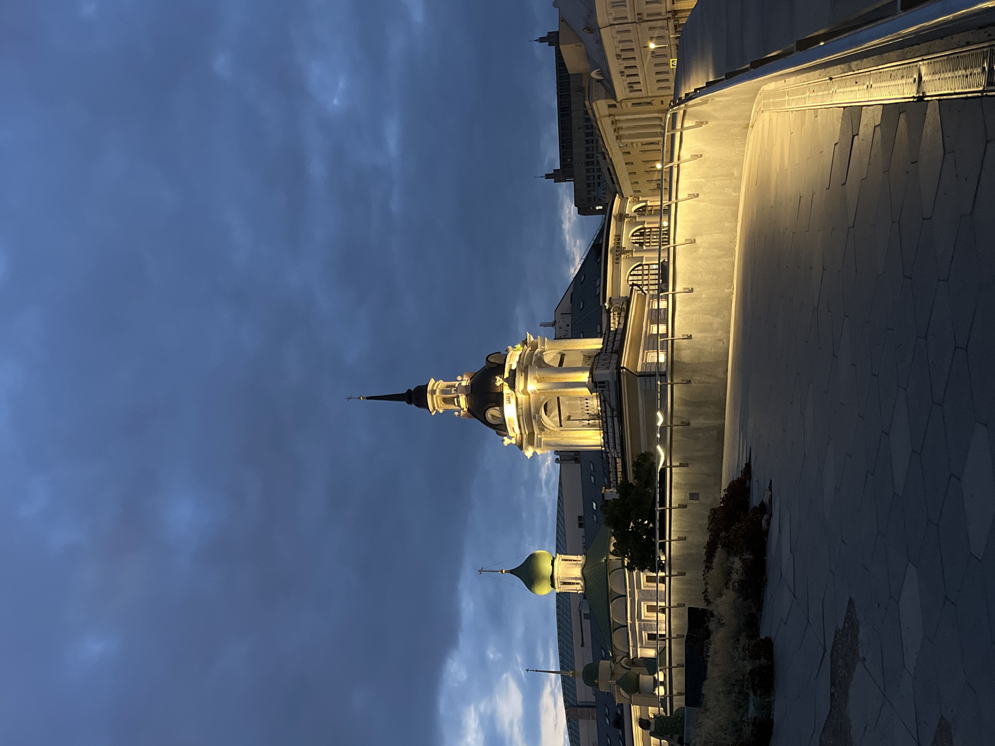 The image depicts a view of Zaryadye Park in Moscow, Russia, at dusk. The sky is a deep blue with dramatic, heavy clouds. Illuminated buildings with distinct architectural styles are visible, including a building with a central, illuminated dome topped with a spire and cross, and another building with a green dome and a cross. These structures are part of historic or religious edifices. A walkway with distinctive geometric paving stones leads into the foreground. The walkway is bordered by a light-colored, curved wall that is illuminated from above, casting a warm glow. A metal railing runs along the edge of the walkway. In the background, other buildings with classical architectural features, such as columns and multiple windows, are visible, also illuminated. The scene is devoid of any visible individuals. The overall impression is one of a twilight cityscape, highlighting the illuminated architecture against a darkening sky.
