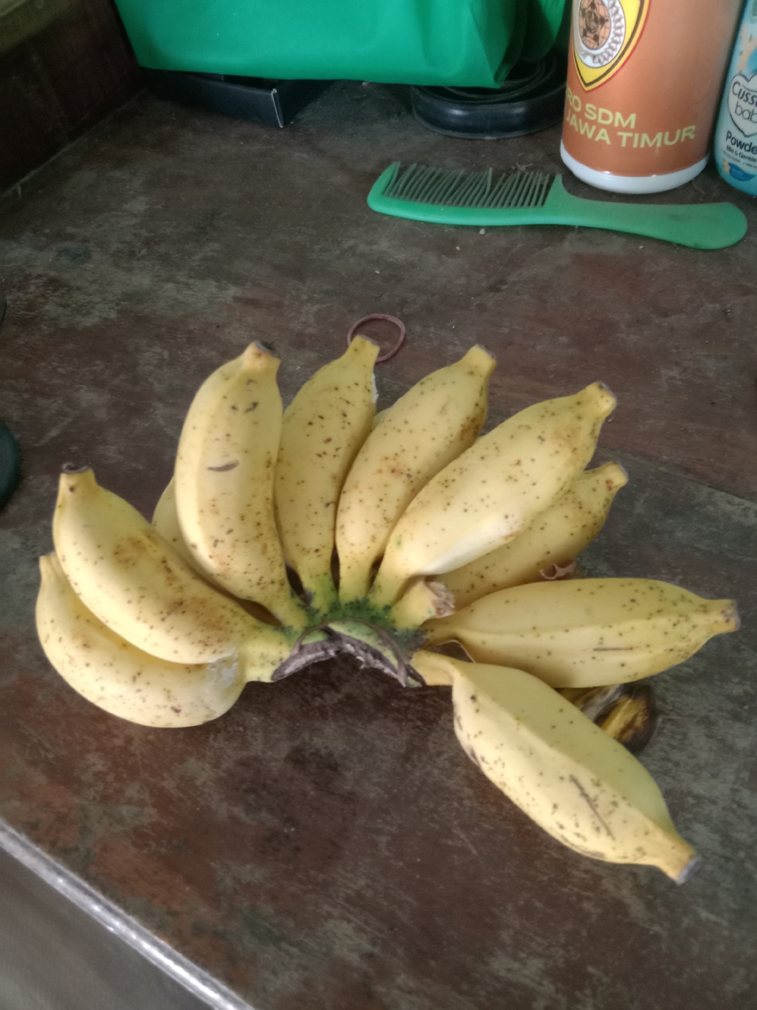 A bunch of yellow bananas, some with brown spots, are laid out on a dark, textured wooden surface. The bananas are the central focus of the image. In the background, a green object, possibly a cloth or bag, is visible along with a green comb and a container with text that appears to be in Indonesian. The overall setting suggests an indoor environment, likely a kitchen or dining area. The lighting is natural, casting soft shadows. There are no people or animals present in the image.
