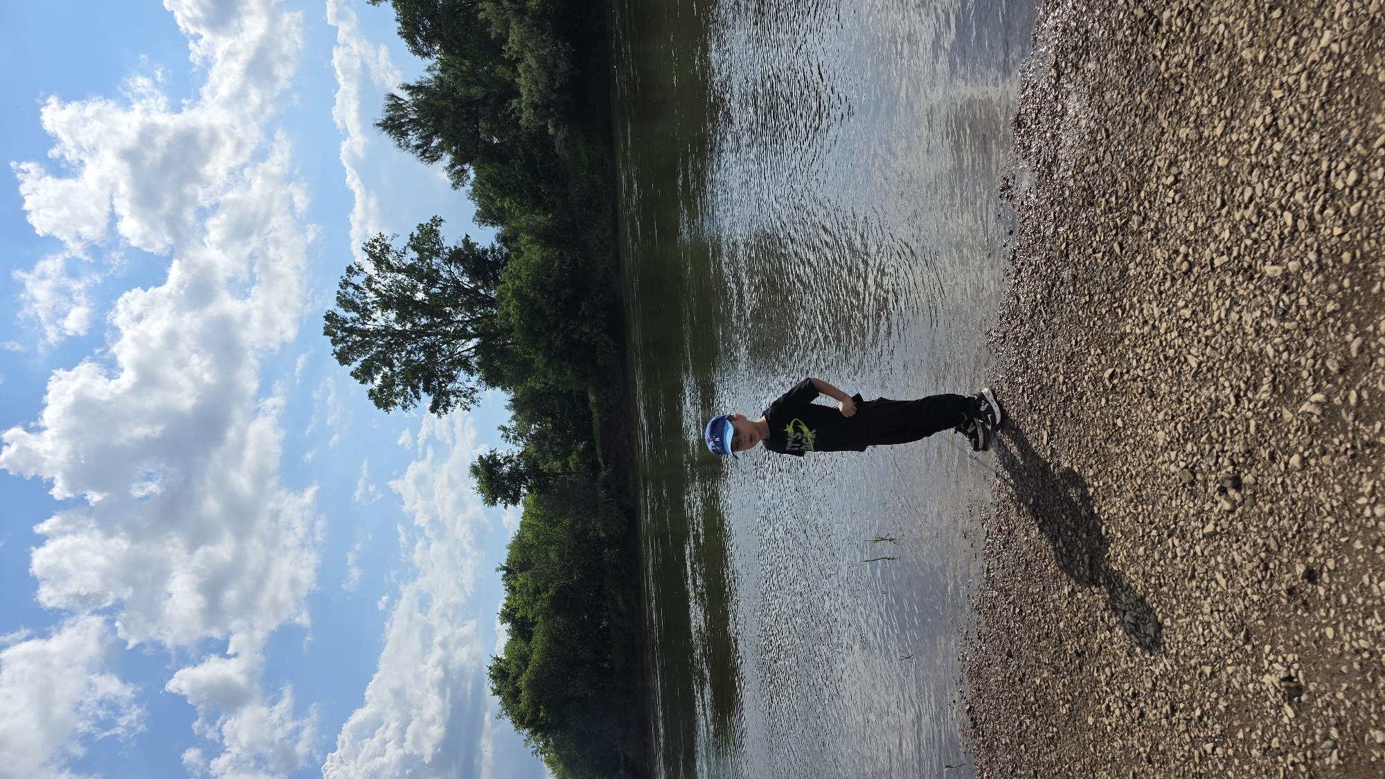 A young boy wearing a blue baseball cap, a black t-shirt, and black pants stands on a rocky shore beside a wide river. The boy is facing slightly away from the camera, with his right hand on his hip. The river's surface is rippled, and a line of green trees forms the opposite bank. The sky above is blue with scattered white clouds. The scene appears to be in an outdoor, natural setting, possibly near the village of Darino, Russia, given the overall landscape and vegetation.