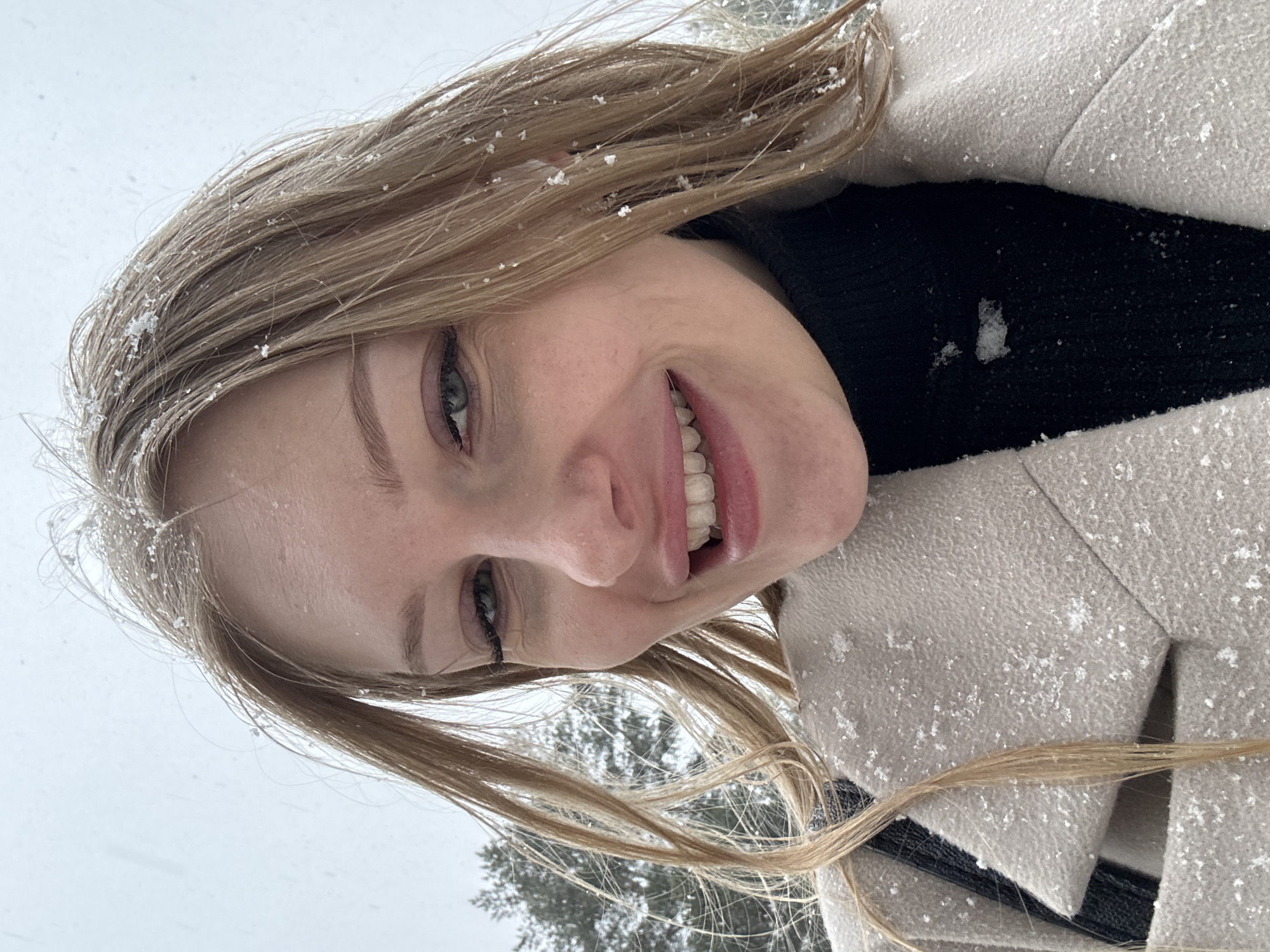 A close-up, angled portrait captures a smiling woman with blonde hair dusted with snow. She wears a black turtleneck under a light-colored coat, also speckled with snowflakes. The background shows a blurred winter landscape, possibly a forest, with snow-covered trees and a gray sky. While the location is reportedly Perm, Russia, specific details confirming this are absent from the visual content.