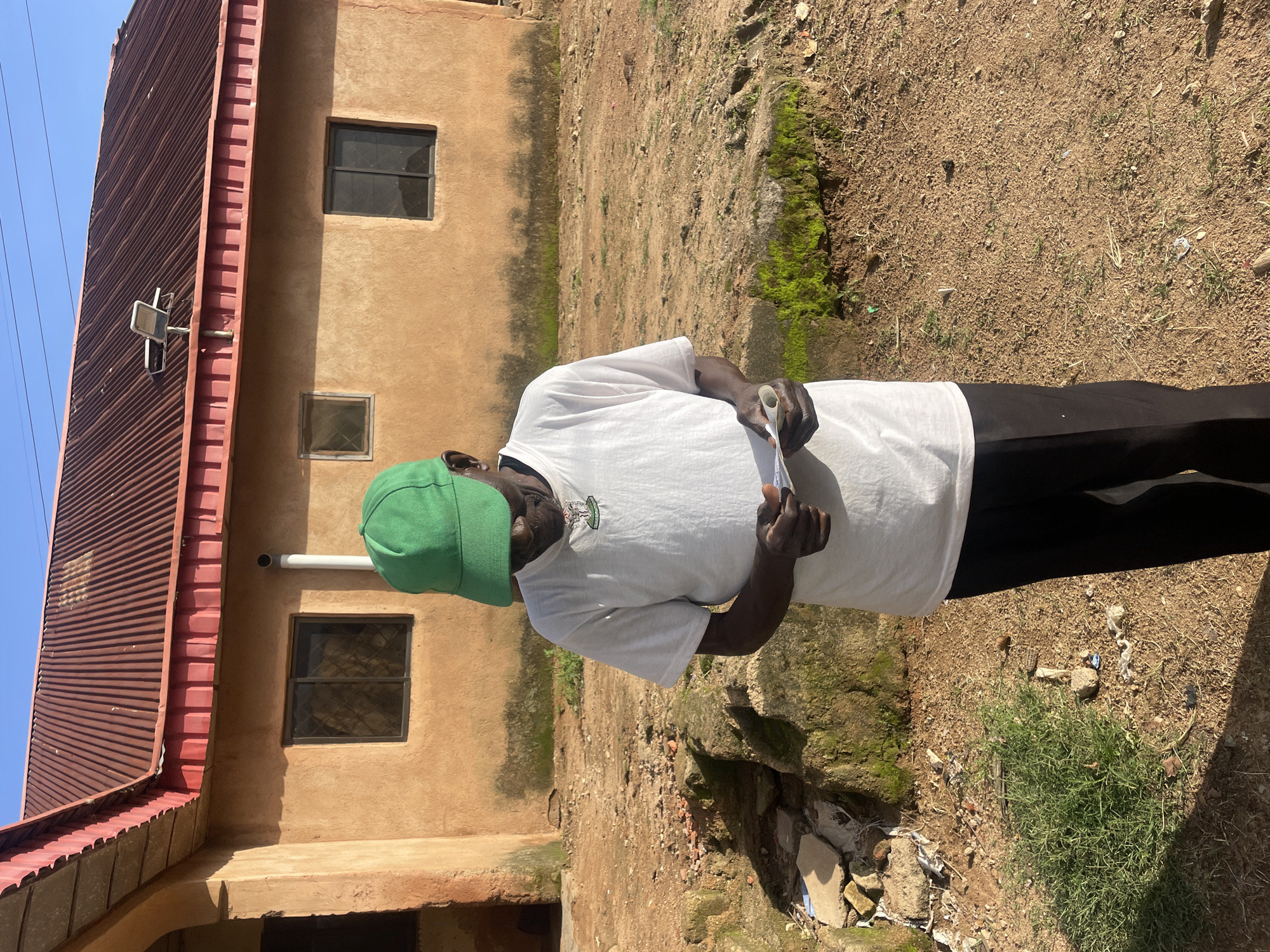 An elderly Black man wearing a white t-shirt and black pants stands outside a tan, single-story building with a red, corrugated metal roof. He is wearing a green baseball cap and looking down at a small piece of paper in his hands. The ground in front of the building is dirt and gravel with scattered debris, including rocks, broken concrete, and plastic. There is a patch of green grass to the left of the man. The building appears to have at least two windows, and a solar panel is visible on the roof. The scene is illuminated by natural sunlight.