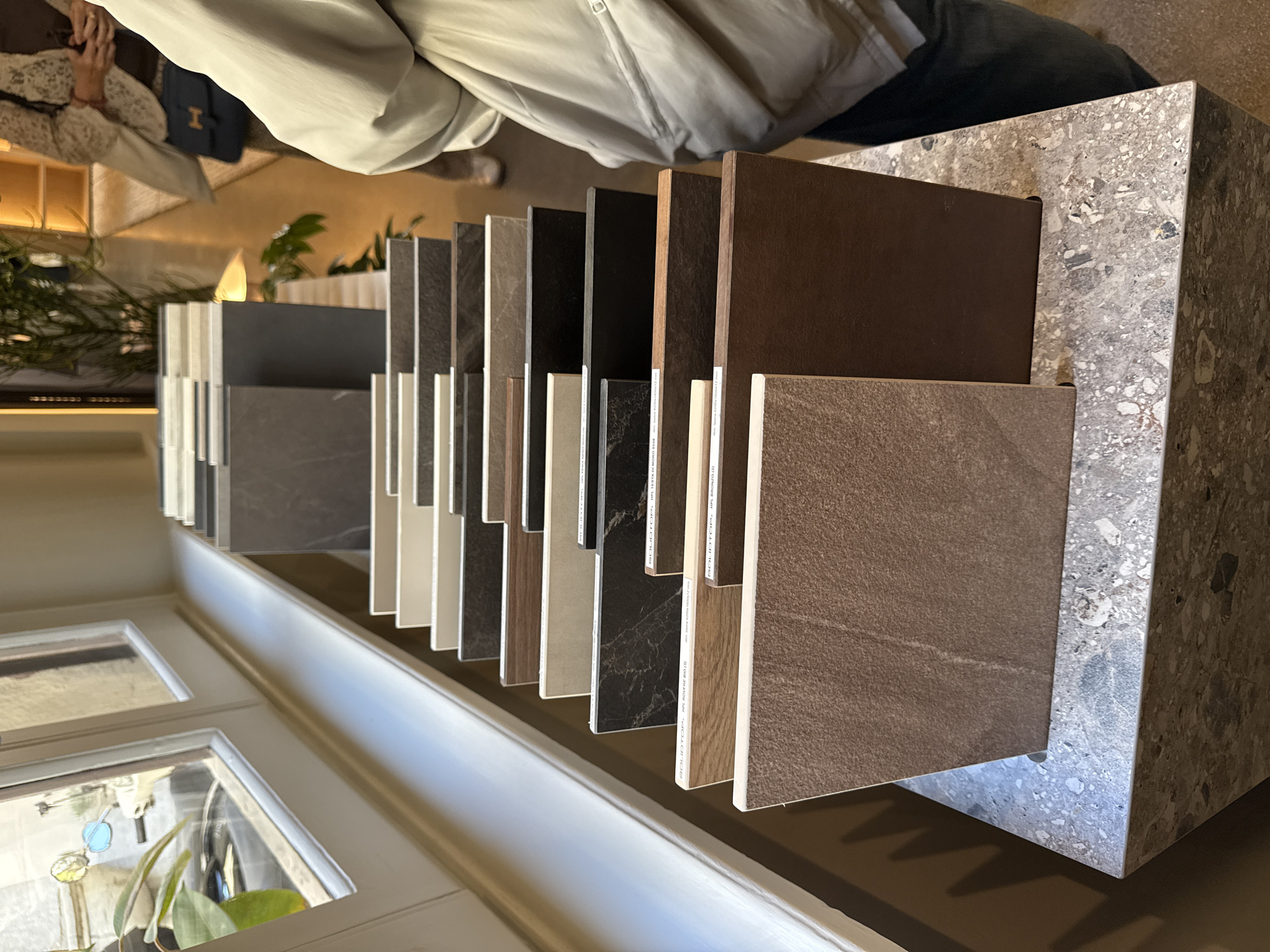 The image captures an indoor scene, likely a design showroom or studio in Milan, Italy, focusing on an extensive display of tile samples.
**Subjects:**
The primary subjects are numerous rectangular tile samples of varying colors and textures, arranged upright in a long, linear display. These samples showcase shades of brown, black, various grays, white, and beige, with finishes mimicking natural stone, concrete, and wood grain. They are held in a custom display unit constructed from a light gray material flecked with darker and lighter aggregates, resembling terrazzo.
In the background, a person is partially visible, wearing a light-colored, possibly beige or cream, long-sleeved jacket. Their hands are obscured, but they appear to be looking down. A dark blue or black bag with a distinctive gold "H" logo (suggesting Hermès) rests near them. Green foliage from plants is visible in the background, and on the far left, a window or door with multiple glass panes shows a glimpse of outdoor light and more plants.
**Activity or event taking place:**
The scene depicts a product showcase or an interior design consultation, where potential clients can view and select different tile materials.
**Notable details:**
The tile samples are neatly organized, allowing for easy comparison of their diverse patterns and finishes. The display stand itself is a key design element, featuring a textured, composite material. The high-end branded bag in the background suggests a sophisticated environment. While it's an indoor setting, the light through the window suggests daytime. The overall aesthetic points to a contemporary and design-conscious space.
**Visible text:**
Labels on several tile samples include brand and product information. Identifiable text fragments are "SOLIDTOP.", "MFL TERRA D", "MFL BRONZO EB", "GRANITO FIESTA DI MONZA R38", and a dimension "4800 x 900 x 12 mm", indicating the size of some of the large format tiles.