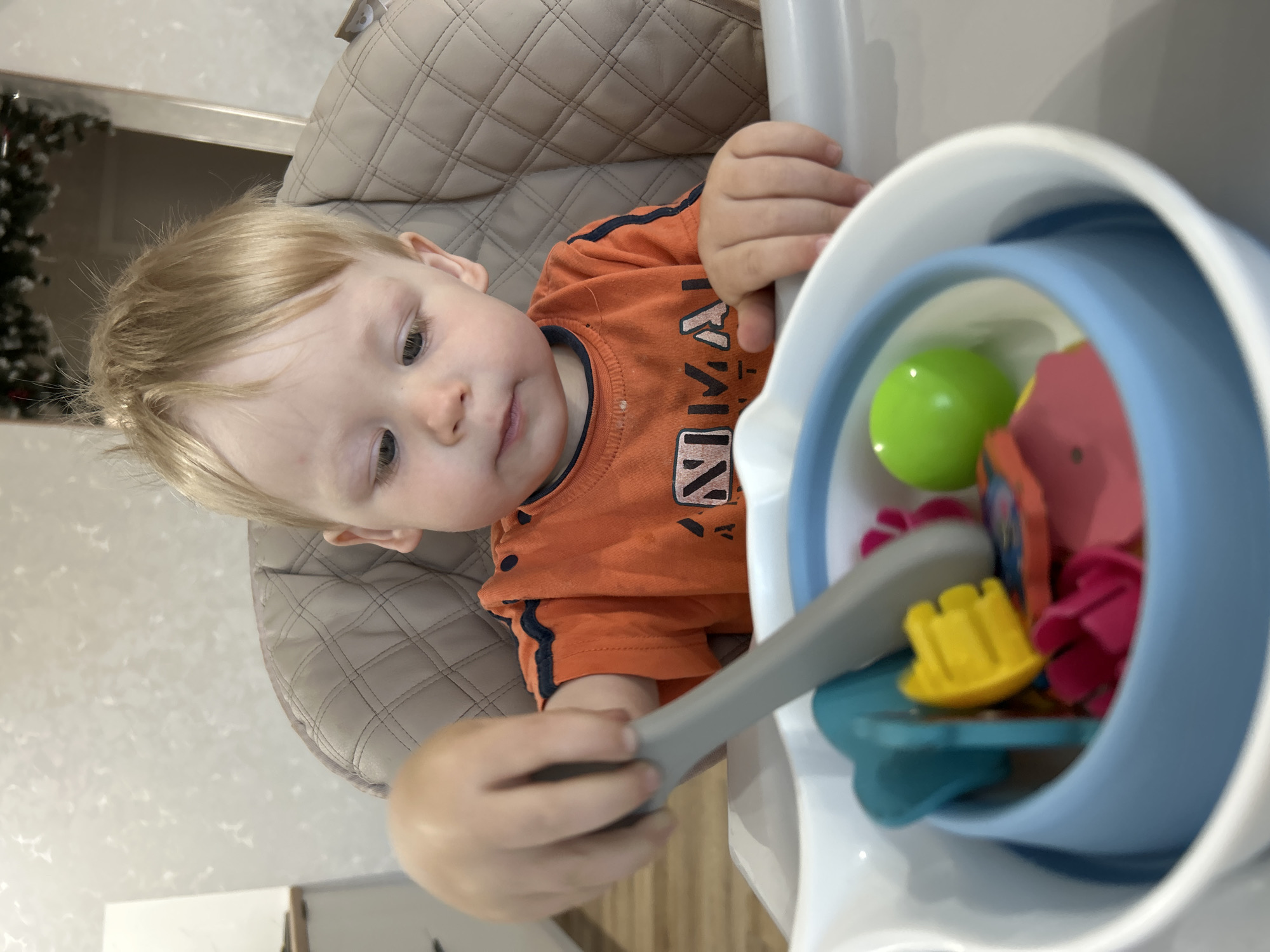 A young child with blond hair sits in a high chair, playing with a toy set. The child is wearing an orange shirt with blue trim. The toy set is in a blue bowl, and includes colorful plastic shapes and a green ball. The high chair is beige and appears to be made of padded material. The background is blurred, but suggests an indoor setting with a wall and a door. This image was taken in Chaikovsky, Russia.