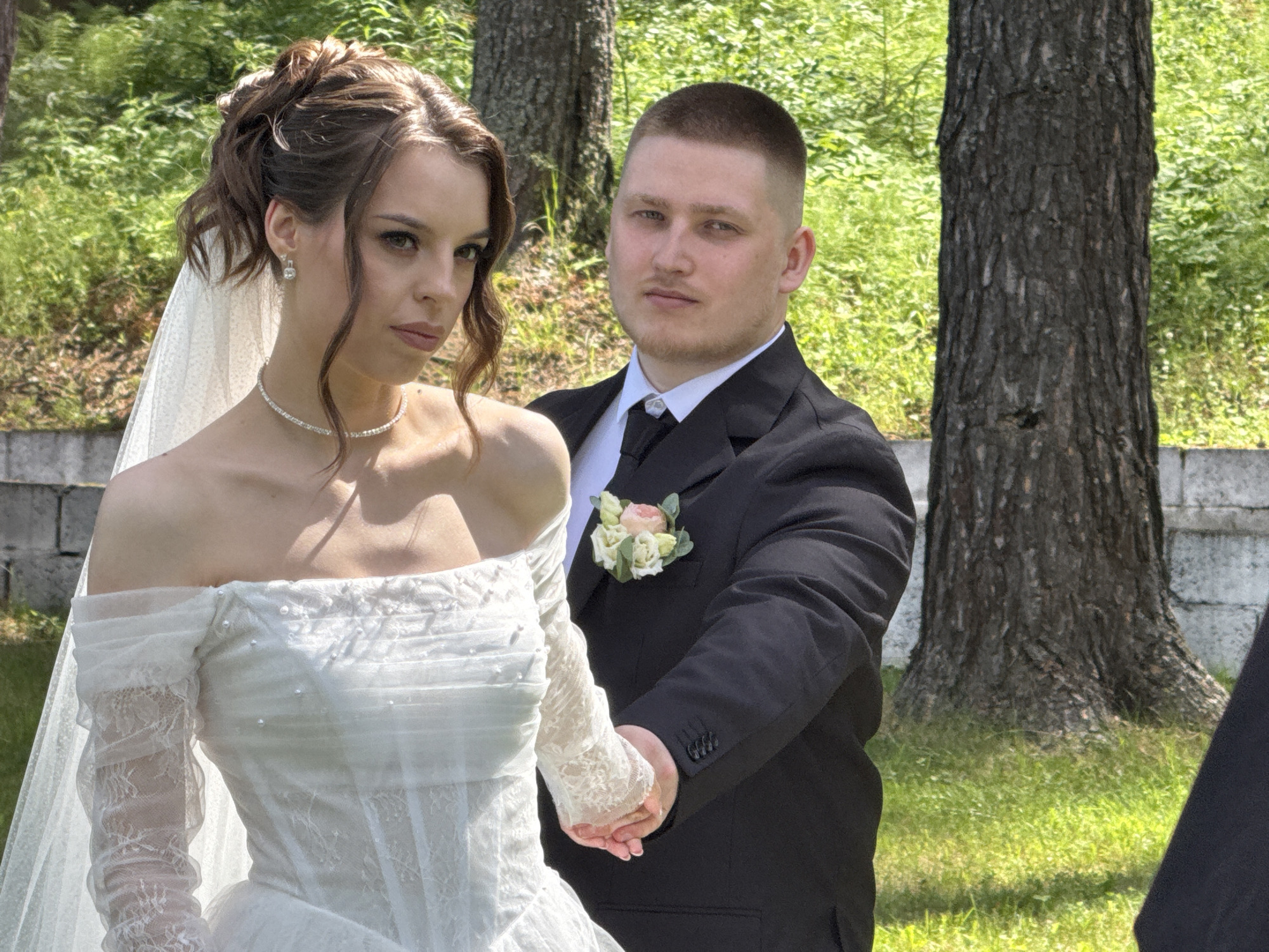 A medium shot captures a wedding portrait of a young couple standing outdoors, with a lush green background of trees and foliage. The bride, on the left, is wearing an off-the-shoulder white wedding dress with lace detailing and off-the-shoulder sleeves. She has her hair styled up, adorned with a veil, and is wearing a delicate diamond necklace and earrings. She is looking directly at the camera with a serious expression. The groom, on the right, is in a black suit with a white shirt and black tie. He has a buzz cut and is looking towards the bride. He is holding her hand, and a boutonnière with light pink and white flowers is pinned to his lapel. The environment suggests a park or garden setting, likely in Votkinsk, Russia, given the presence of birch trees common to the region. The light indicates it is daytime, possibly a sunny day, given the shadows.