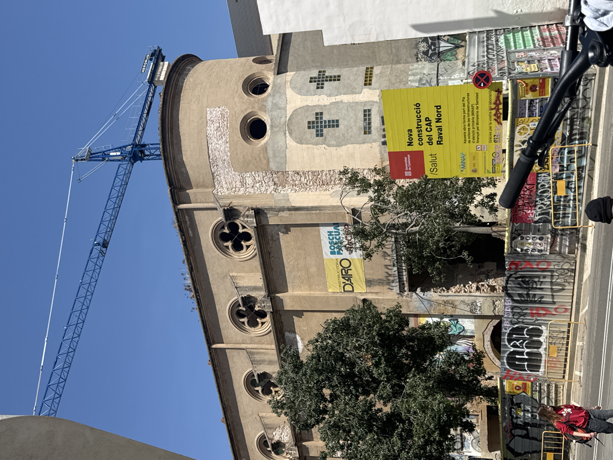 An image captures a construction site in Barcelona, Spain, featuring an ornate, possibly former institutional building or church undergoing renovation. The building's facade is a light brown, with multiple round windows, some designed with quatrefoil patterns, and others as simple circular openings. Higher up, there are two larger circular openings and rectangular windows arranged in distinct cross shapes. Parts of the facade show exposed stonework and signs of repair.
To the left, a large blue construction crane with "HERGRUS" written on its arm extends into a clear, bright blue sky, indicating ongoing work. Below the crane, scaffolding or protective barriers cover portions of the building's exterior.
In the mid-ground, a prominent yellow sign reads "Nova construcció del CAP Raval Nord" (New construction of the Raval Nord Primary Care Center) and "/Salut," indicating the purpose of the project. Further text on the sign, in Catalan, states "Generalitat de Catalunya," "MINAP," and details that the work is part of a plan to improve primary care infrastructure, funded by the Ministry of Health. It also includes "Gràcies per la vostra col·laboració" (Thanks for your collaboration) and "012." Another yellow banner visible on the building identifies "CONSTRUCTORA D'ARO BOSCH PASCUA."
The foreground features metal construction barriers heavily covered in colorful graffiti. A red no-parking sign is also visible. Partially obscured by the barriers and trees on the right, a person walks, wearing a red shirt with a floral design and dark pants, carrying a bag. The bright lighting suggests it is daytime. The combination of historical architecture, active construction, and urban graffiti provides a snapshot of development in the Raval Nord district of Barcelona.