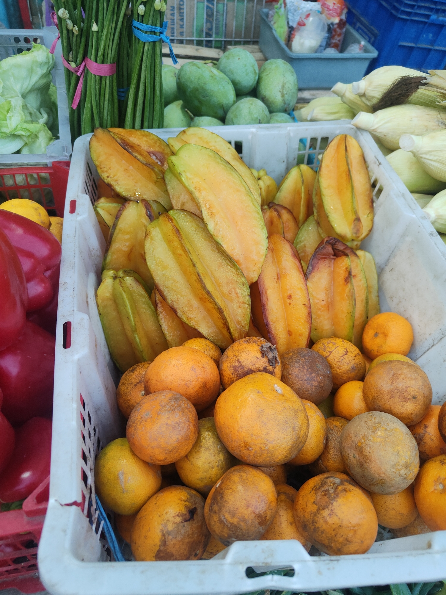 This image shows a close-up of a fruit stand in Kauman, Indonesia. In the foreground, a white plastic crate is filled with starfruit and oranges. The starfruit is cut in half, revealing its star shape and yellow flesh. The oranges are whole and have rough, bumpy skins. Behind the crate of fruit, other produce is visible, including green lettuce, red bell peppers, and corn on the cob. The overall scene is a vibrant display of fresh produce, typical of a local market.