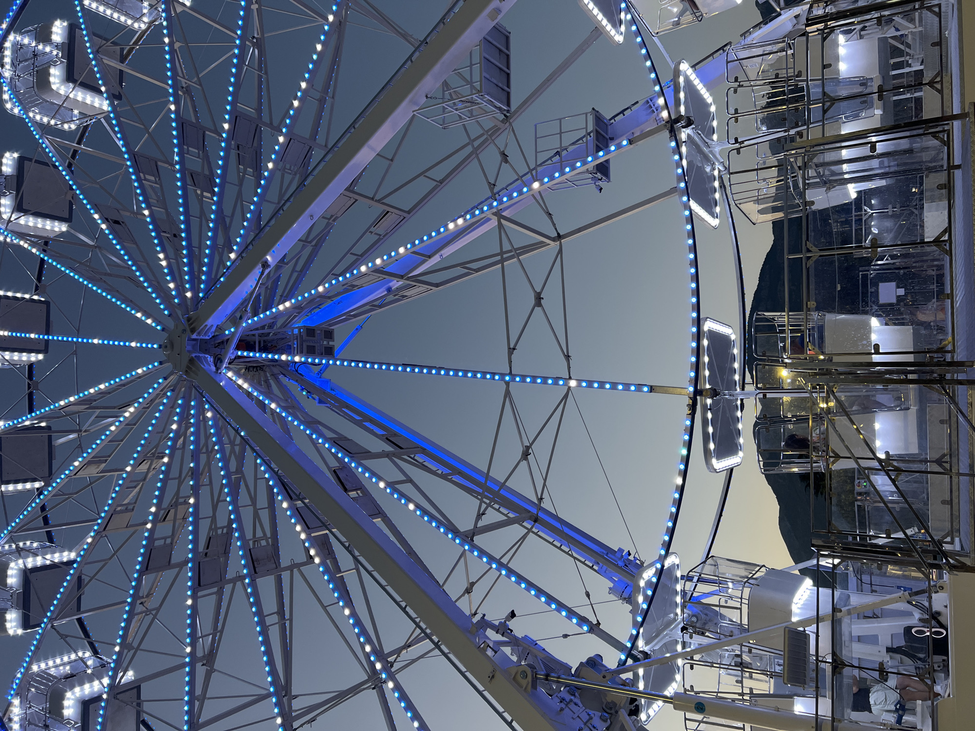 The image is a close-up, low-angle shot of a Ferris wheel illuminated with blue and white lights against a twilight sky. The Ferris wheel is the dominant subject, with its intricate metal structure and numerous spokes extending outwards. Several gondolas, enclosed with clear panels and metal railings, are visible at the bottom, some with people inside. In the background, silhouetted mountains are visible against the fading light. The setting appears to be an amusement park or fairground. While the exact location is not specified in the image, the presence of mountains suggests a scenic, potentially coastal area.