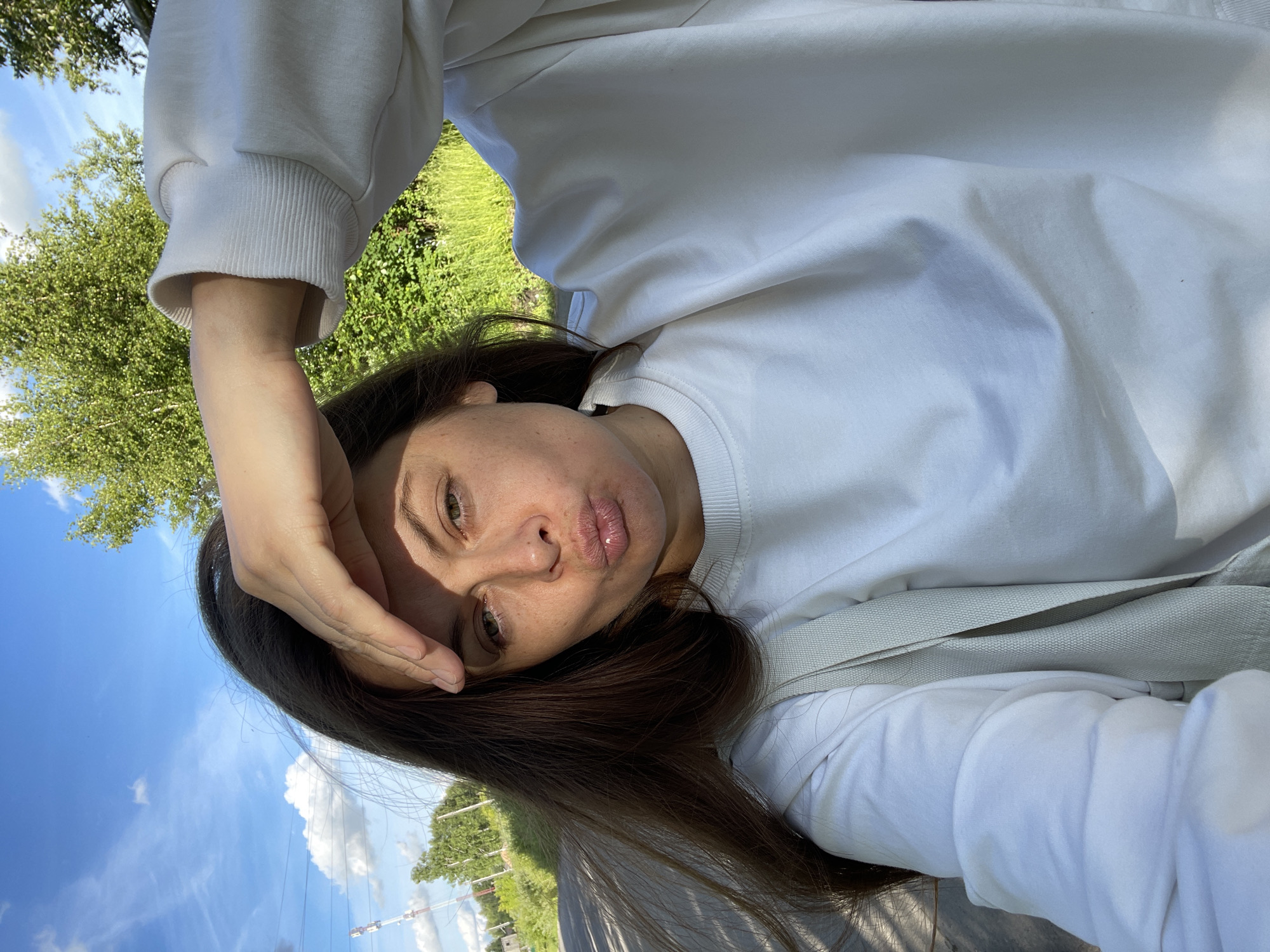 A young woman with long brown hair, wearing a white sweatshirt and a light-colored strap across her chest, is taking a selfie outdoors on a sunny day. Her right hand is raised to shield her eyes from the sun, and her lips are pursed. The background consists of a blue sky with scattered clouds, green trees, and what appears to be a road or path. There are also some structures, possibly utility poles, visible in the distance. The overall impression is that the photograph was taken in a rural or semi-rural setting, potentially on the outskirts of the city of Zarechny, Russia.