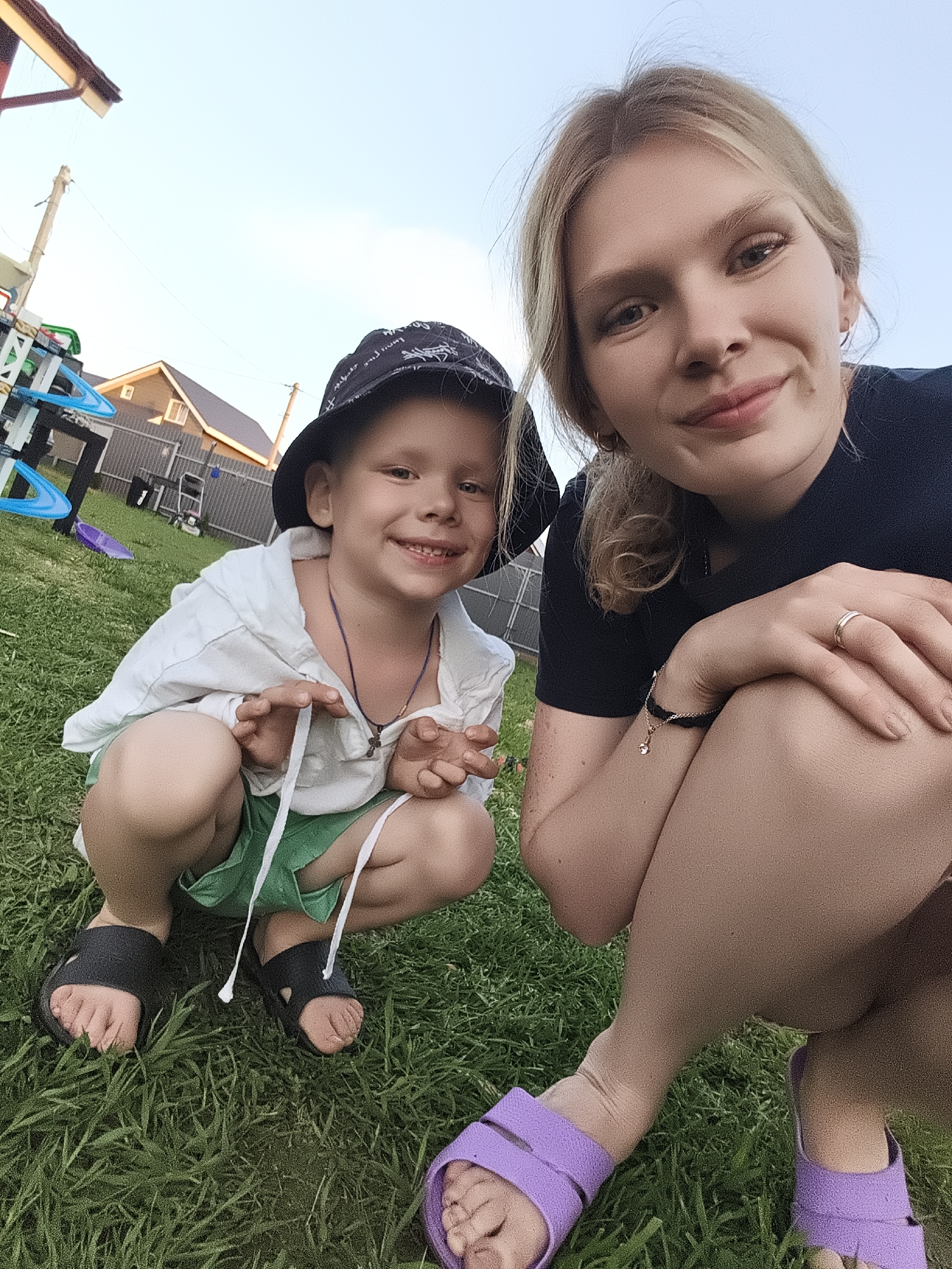 The image shows a blonde woman and a young boy crouching on a lawn in Русский Вожой, Russia. The boy wears a patterned bucket hat, a white hooded shirt, green shorts, and black sandals. He is smiling and appears to be interacting with someone out of frame. The woman, wearing a black top and purple sandals, is also smiling and has her arm resting on her knee. The background includes a house, a playset, and a fence. The scene is set outdoors during daylight hours.