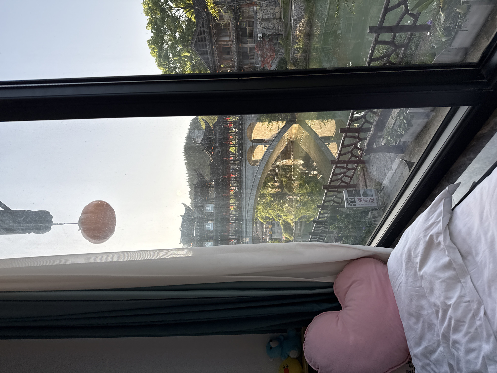 A view through a window shows a city scene in Furong, China. The window's dark frame surrounds a landscape that includes a stone arch bridge and traditional Chinese buildings. The bridge is reflected in the water below. The view is partly obscured by a white curtain, a pink heart-shaped pillow, and a white pillow on a bed. A hanging decoration is visible in the upper left of the window.