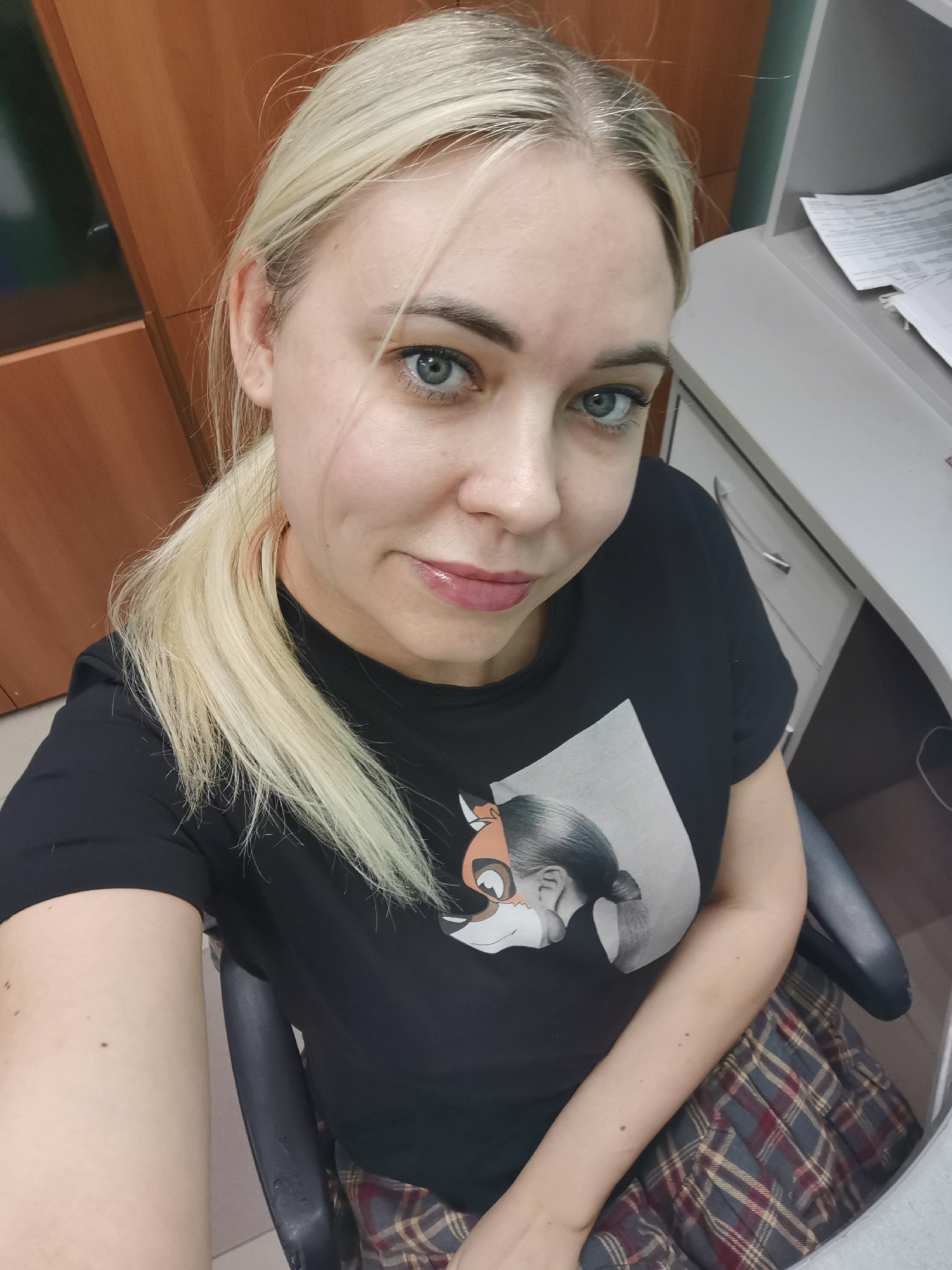 A fair-skinned woman with blonde hair pulled back into a ponytail is seated in an office chair. She is wearing a black t-shirt with a graphic of a woman and a fox on it, and a plaid skirt. The background includes wooden cabinets on the left, a gray desk with paperwork on the right, and a drawer. The location is Chaikovsky, Russia. The woman appears to be taking a selfie.