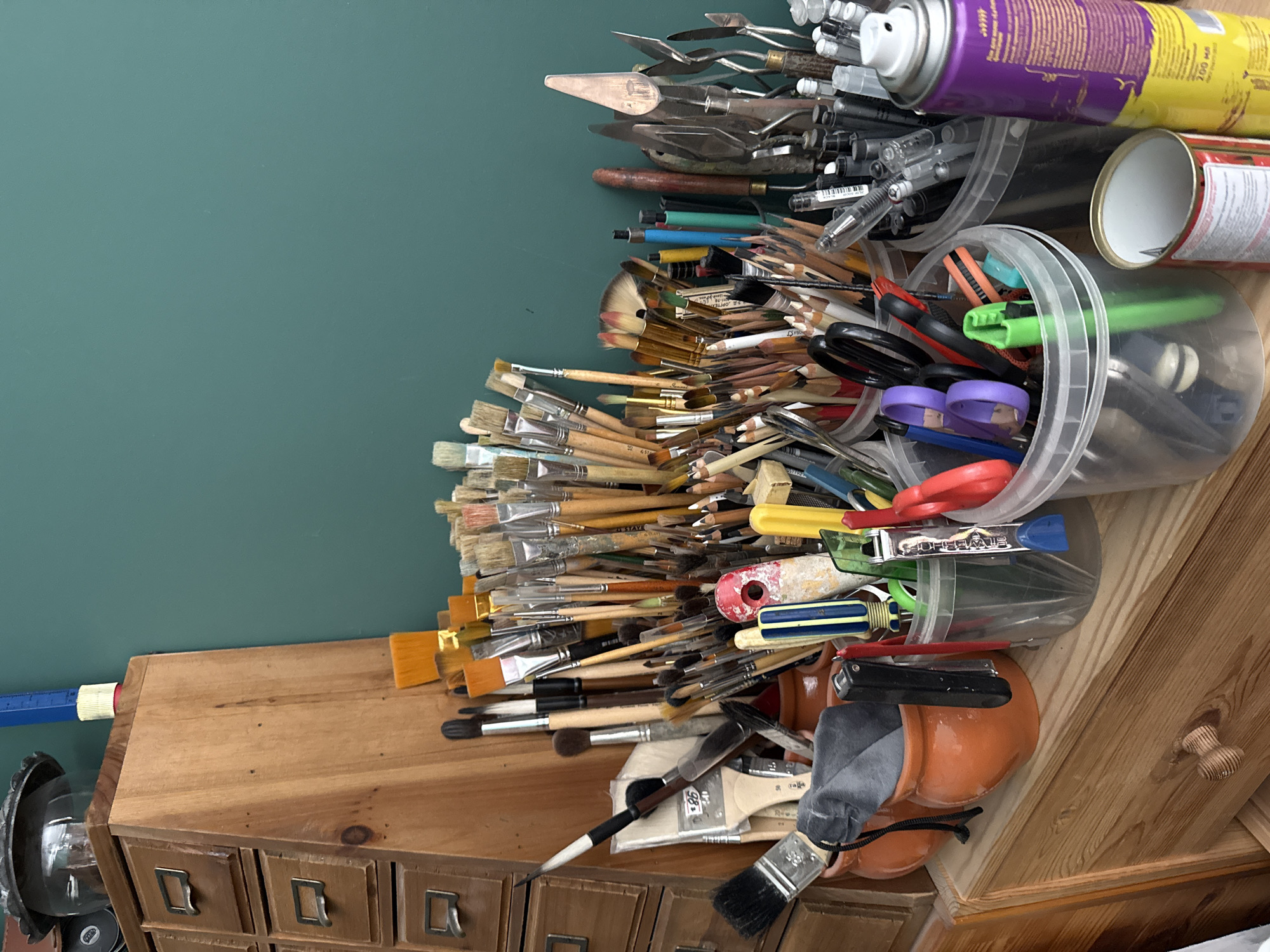 The image shows an artist's workstation, likely in Votkinsk, Russia, with various art supplies neatly organized. The scene is dominated by a wooden cabinet with multiple drawers, filled with an assortment of paintbrushes, pencils, palette knives, scissors, and spray cans. The brushes vary in size and bristle type, indicating a range of artistic techniques. Pencils are sharpened and arranged in containers, alongside colorful crayons and markers. Palette knives are presented in different shapes and sizes, suggesting various applications in painting. Scissors and other cutting tools are also visible. A can of spray paint with purple and yellow labeling stands prominently, hinting at mixed-media art. The background features a teal-colored wall, providing a contrasting backdrop to the wooden furniture and art supplies. The overall impression is one of a well-equipped and organized creative space.