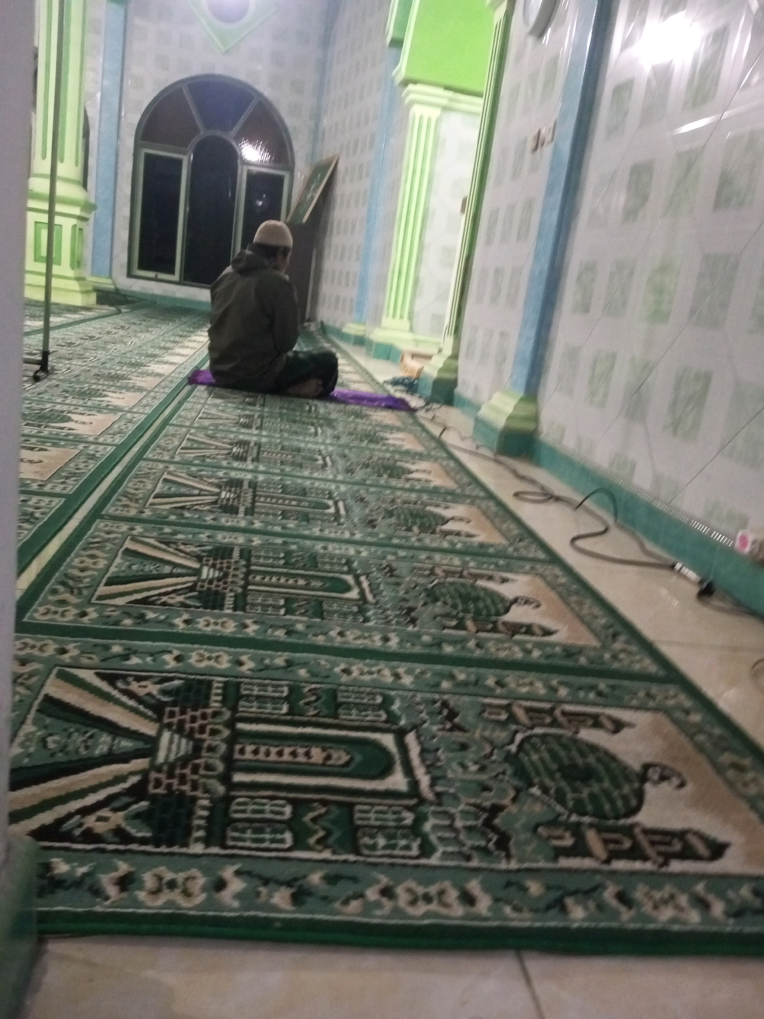 A person wearing a white cap and a dark green hoodie is seated on a purple prayer mat on a green carpet with a repeating pattern in a room with tiled walls. The person is facing away from the camera, towards a large arched window. The room appears to be a place of worship, likely a mosque, given the prayer mat and carpet. The walls are adorned with light-colored tiles, and there are green pillars and trim. The floor is made of tiled material. The image is dimly lit, suggesting it was taken indoors with artificial lighting or at a time of day with limited natural light. The location is identified as Andongsili, Indonesia.