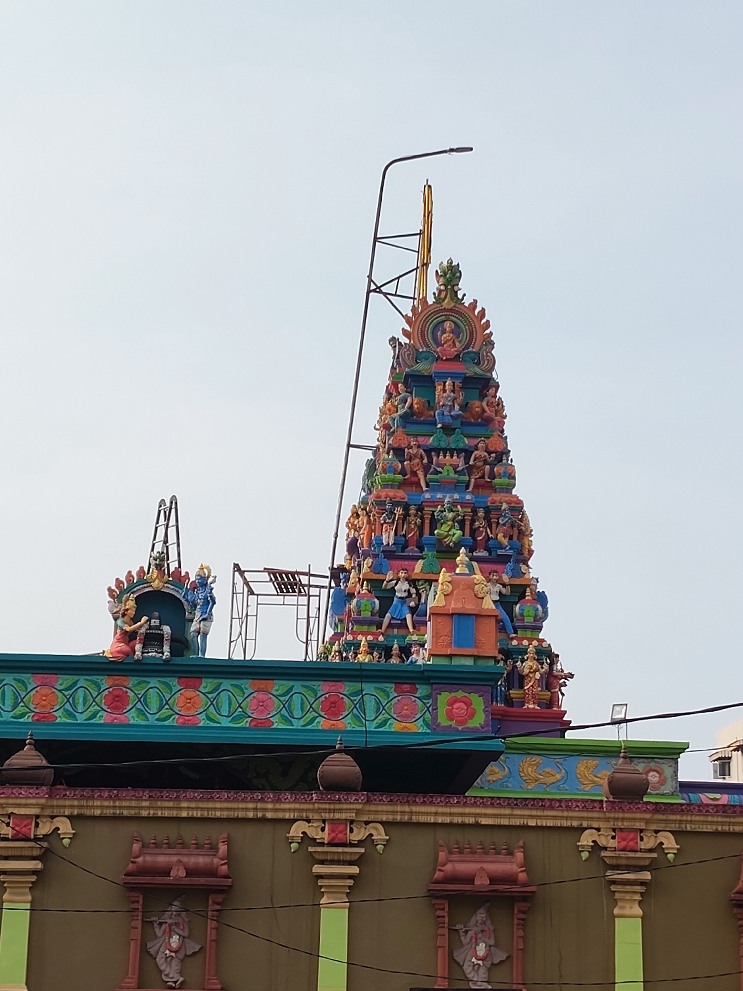 The image shows a colorful Hindu temple in Medan, Indonesia. The temple features a tall, tiered tower adorned with numerous statues of deities and mythical figures. The foreground shows the upper part of the temple building with decorative friezes and architectural details, including pillars and ornamental urns. A street lamp and some construction scaffolding are also visible near the temple's pinnacle. The sky is overcast and pale.