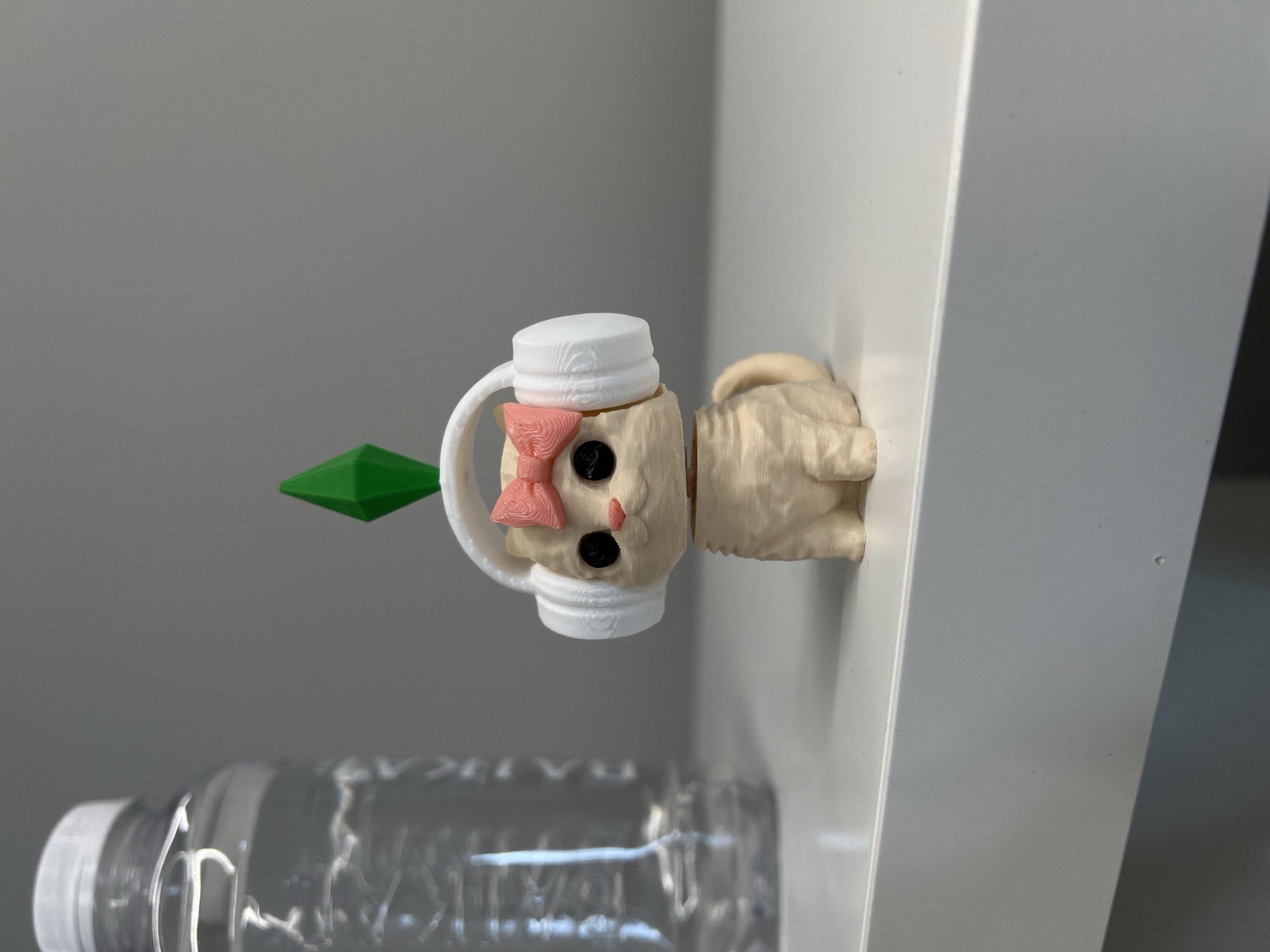 The image displays a light-colored, possibly 3D-printed, figurine resembling a stylized creature, positioned horizontally. The figurine has a distinct head featuring two black, round eyes, a textured pink bow on top, and a white, ridged cap or hat. It holds a bright green, spiky, multi-faceted object in what appears to be its left hand. The figurine's body extends to the right, where it is attached to or resting against a white, vertical panel. This panel forms a corner with another white surface on the right side of the frame. In the background, a plain gray wall is visible. In the lower left of the image, a clear plastic water bottle with a white cap lies horizontally on a surface. Indistinct, blurry text or branding can be seen through the plastic of the water bottle. The scene suggests an indoor setting, likely a room with minimalist decor. The figurine appears to be a static decorative item or a mounted toy. No specific time of day, weather, or emotions are discernible. The location context is Khimki, Russia.