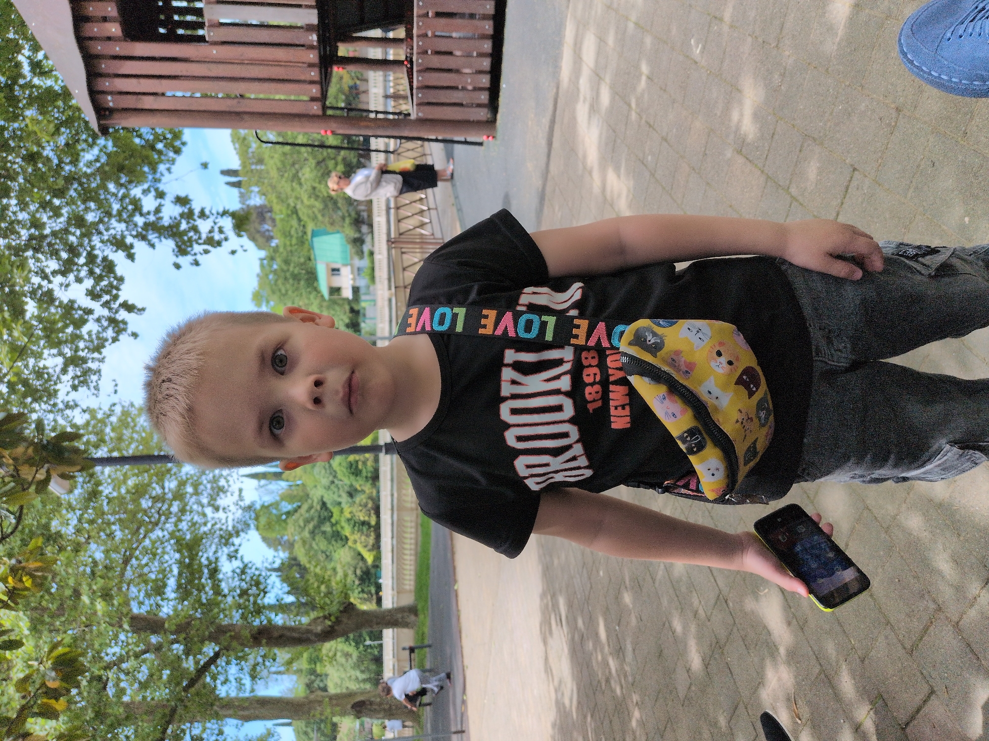 A young boy stands outdoors on a paved area. He wears a black t-shirt with "BROOKLYN" in vertical letters and "LOVE" repeating horizontally across the upper portion. He has a yellow patterned waist bag. He holds a smartphone in his right hand. To the left, trees are visible. In the background, there is a view of greenery and what appears to be a park or recreational area. A partial view of a playground structure is in the upper left corner. In the distance, a person is visible standing near a railing and another person is sitting. Based on the location, this could be in the city of Sochi, Russia.