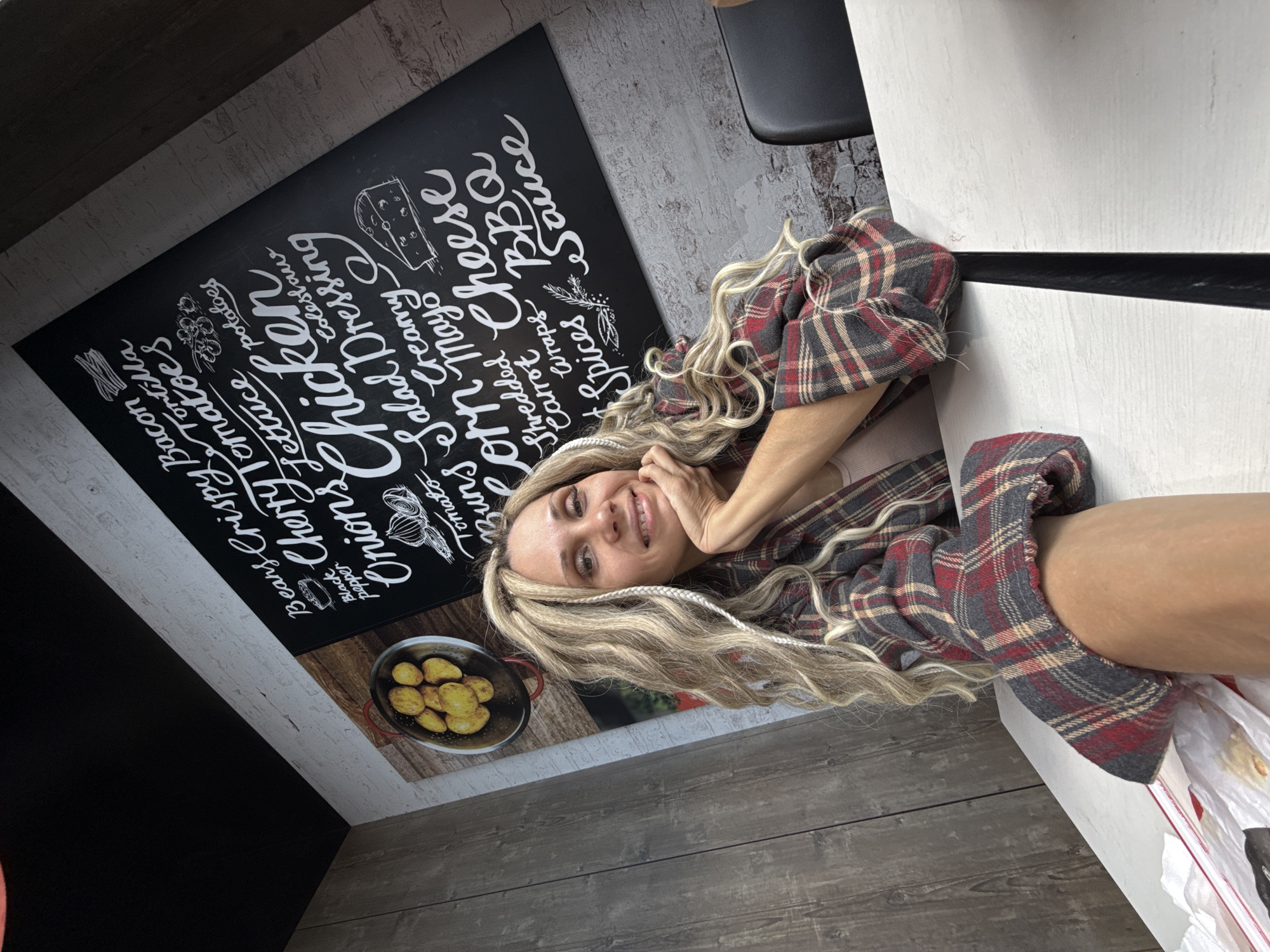 A woman in a plaid shirt is sitting at a table in what appears to be a restaurant or cafe. Behind her is a wall with a chalkboard-style menu listing various food items. The menu includes items such as "Crispy Bacon", "Chicken", "Salad Dressing", "Shredded Cheese", and "Sauce". The woman is smiling and has her hand near her face. The location is Заволжье, Russia.