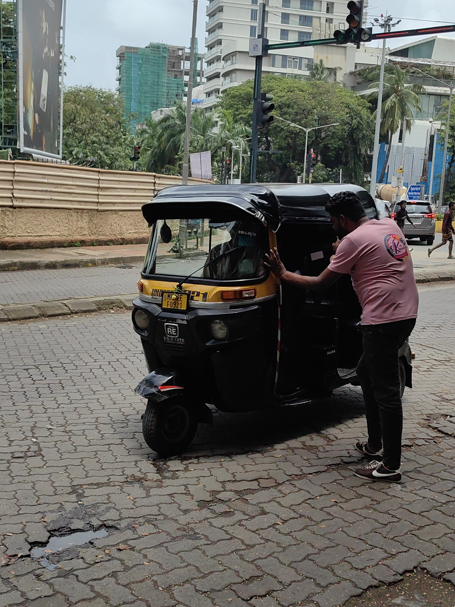 A man in a pink t-shirt and dark pants stands next to a black and yellow auto-rickshaw on a paved street in Mumbai, India. The man's left hand is on the auto-rickshaw's door. The auto-rickshaw has a yellow license plate with "MH 01 E F0921" written on it. Behind the auto-rickshaw, a silver car and two pedestrians are visible on the street. In the background, there are buildings, trees, and a billboard. The sky is overcast. The street is paved with interlocking concrete blocks, and there are several potholes and puddles. The scene suggests an urban street environment in Mumbai.