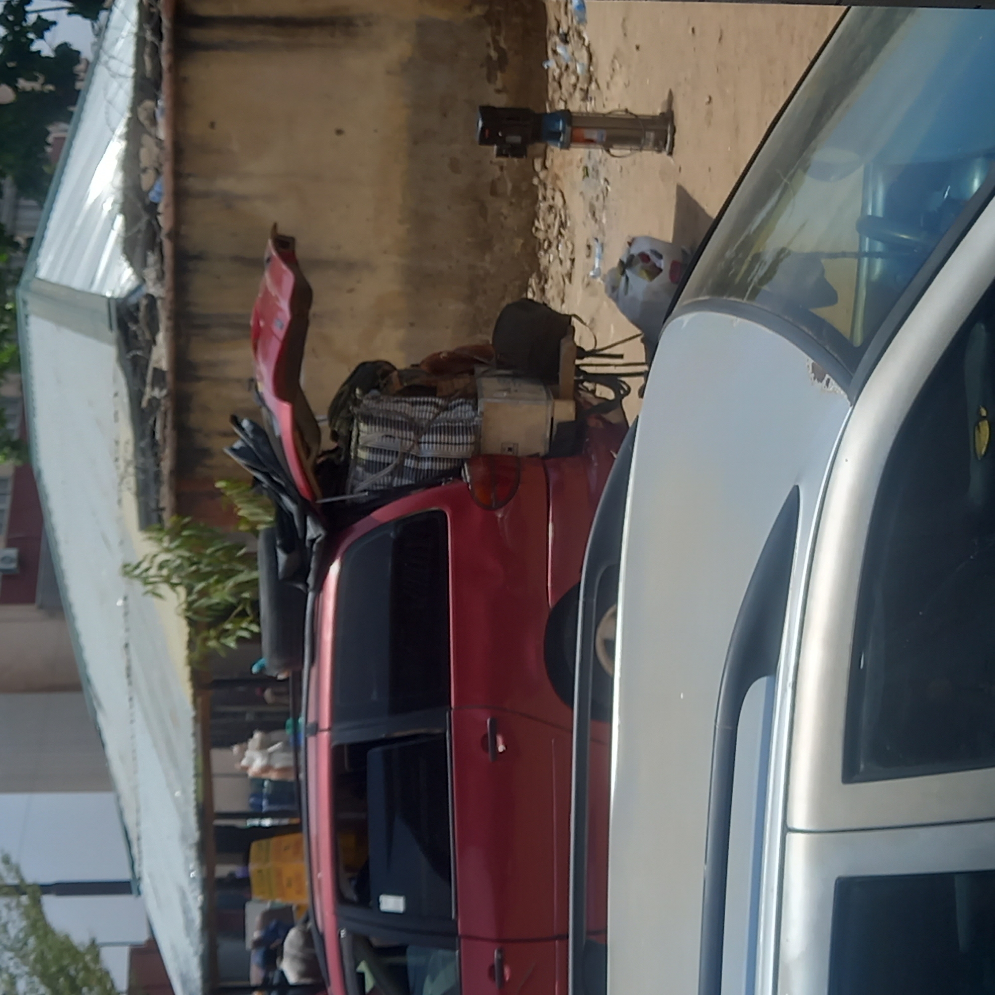 The image shows a red van heavily loaded with goods parked next to a silver car. Debris litters the ground. The setting appears to be an urban environment, possibly Dadi Riba, Nigeria, based on the request. There are buildings in the background and possible individuals on the left. The photograph suggests an informal economic activity, where vehicles are used to transport goods.