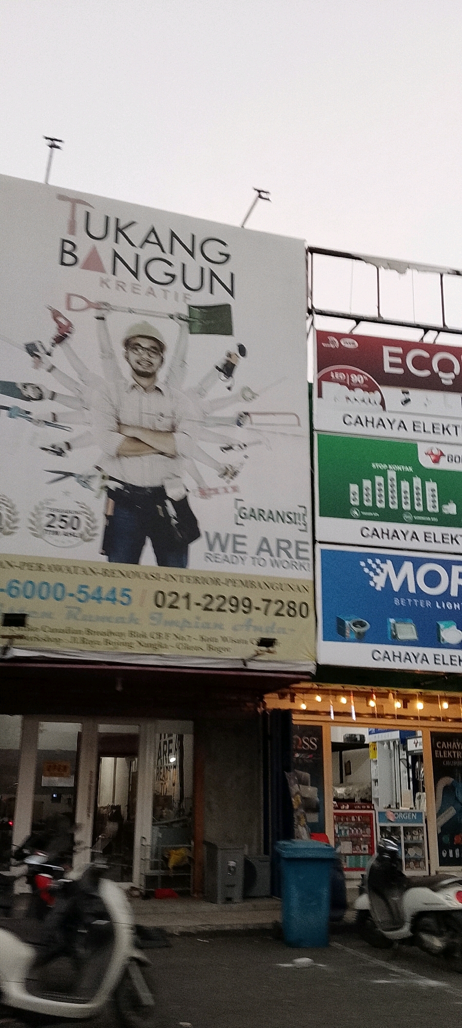 The image shows a street scene in Cileungsi, Indonesia, with a focus on a large billboard and shop fronts. The billboard advertises "TUKANG BANGUN KREATIF," which translates to "Creative Construction Worker," and depicts a man with many arms holding various tools. Below this, another section of the billboard shows "ECO" and "CAHAYA ELEKTRIK." Further down, a green sign displays "STOP KONTAK" (power outlet) and "CAHAYA ELEKTRIK," and a blue sign with the word "MOR" and "BETTER LIGHTING" also advertises "CAHAYA ELEKTRIK."
In the foreground, a street with asphalt pavement is visible, with a white scooter on the left and a white and black scooter on the right, both partially blurred due to motion. In front of the shops, a blue trash bin is prominent. The shop on the left has a glass door with signs indicating "ARE AREADY TO." The shop on the right displays various electrical products and has a sign for "JORGEN." The overall scene suggests a commercial area with a focus on construction and electrical supplies. The sky is overcast.