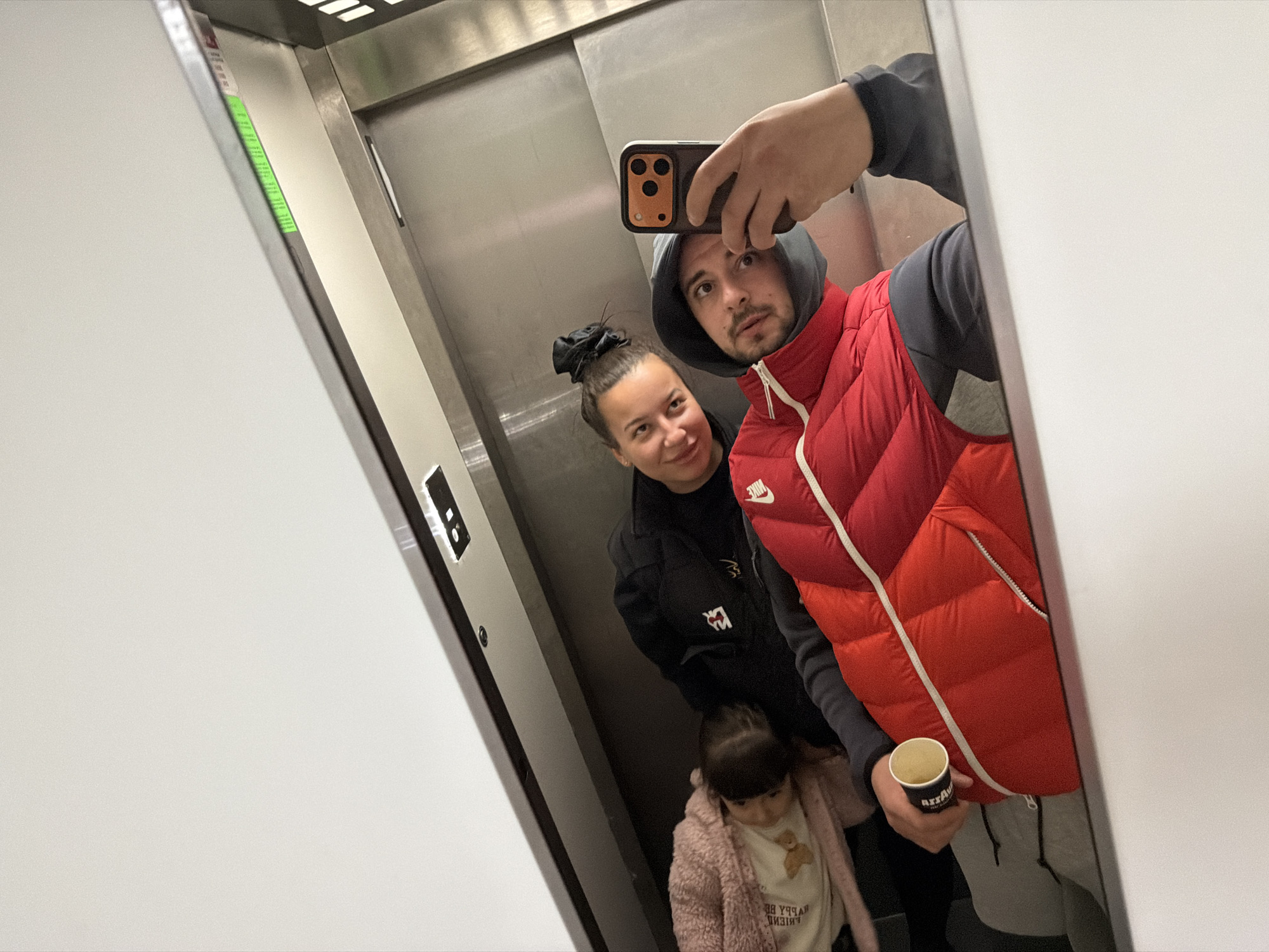 The image appears to be a selfie taken inside an elevator. Three people are visible: a man holding a phone taking the picture, a woman standing behind him, and a young child in the foreground. The man is wearing a red puffer vest over a hooded sweatshirt and holding a coffee cup in his left hand. The woman has her hair up in a bun and is wearing a dark jacket. The child is wearing a pink fuzzy jacket over a white shirt with a teddy bear design. The elevator has metal walls and a control panel visible on the left side. The location in the city of Unknown, Unknown is not able to be determined.