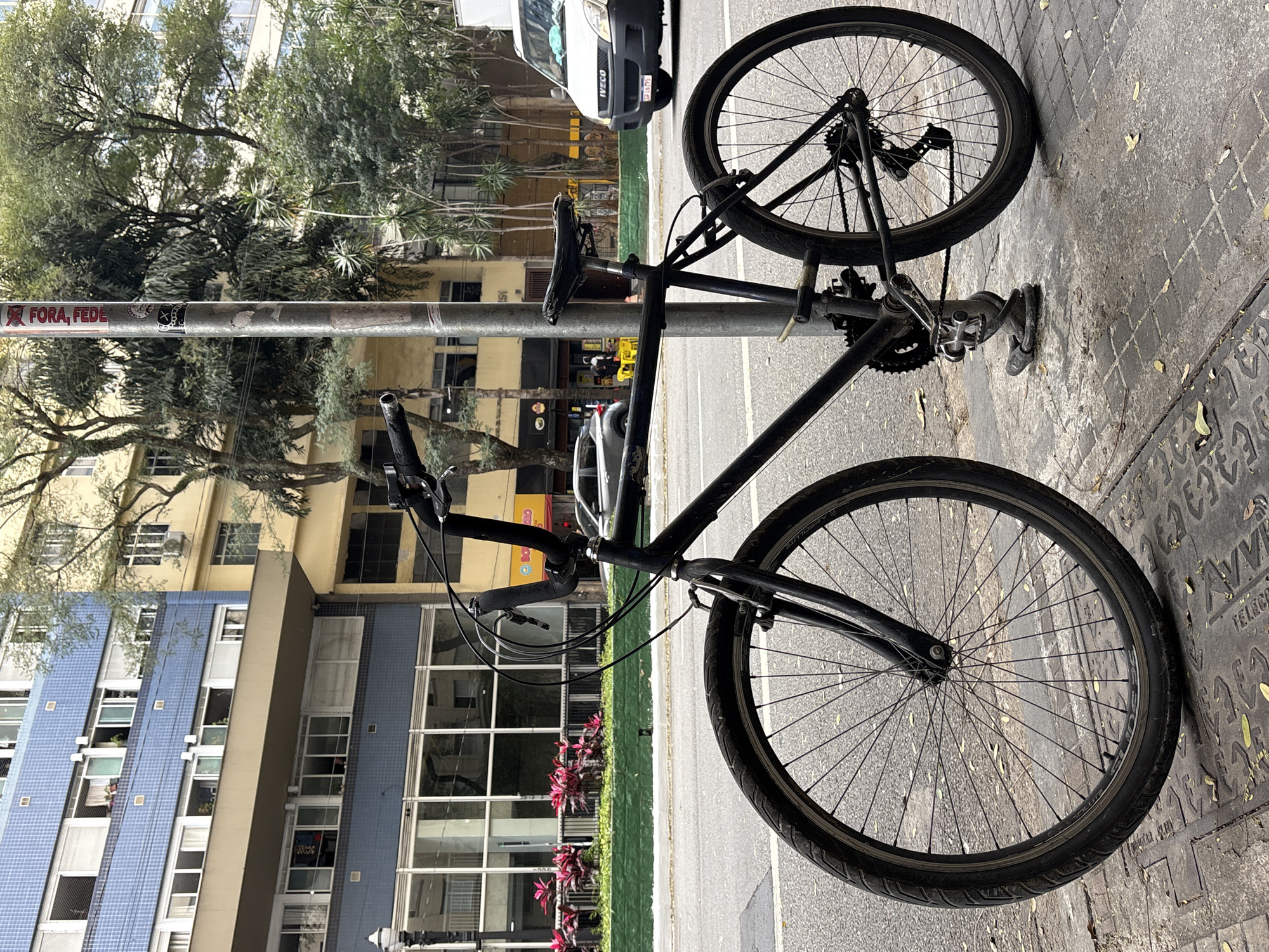 An urban scene in São Paulo, Brazil features a black bicycle locked to a metal pole on a sidewalk. The bicycle is dark-colored, with a visible chain and gears. The pole has several stickers, including one that reads "FORA, FEDE." In the background, several buildings are visible, with a light-colored van parked on the street. The street is paved, and the sidewalk is made of concrete tiles. Trees are present, and the sky appears overcast. The overall impression is of a city street with parked transportation.