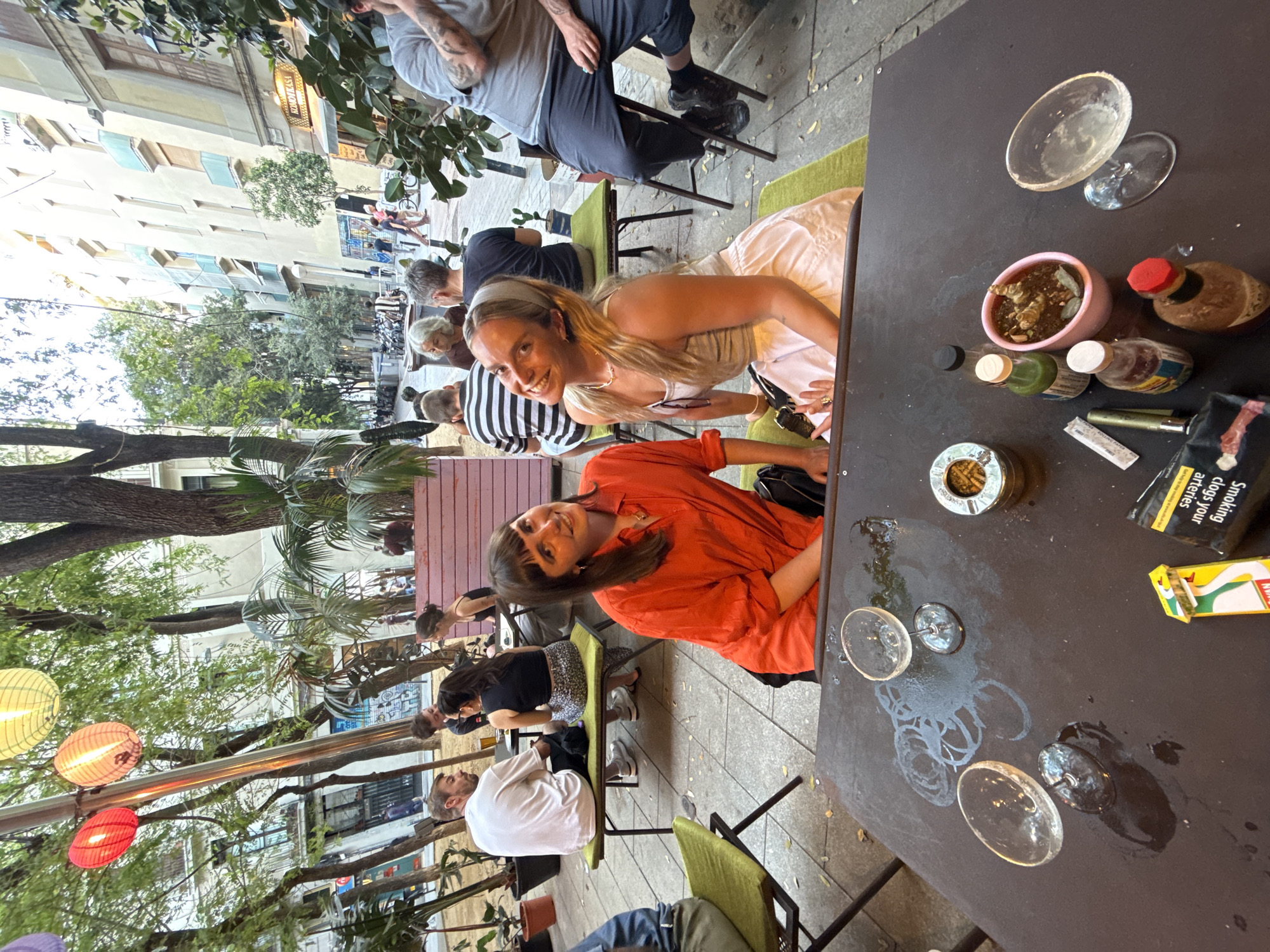 The image captures an outdoor cafe scene in Barcelona, Spain, likely during the daytime or early evening, characterized by natural light filtering through trees. The setting features paved ground, green padded chairs, and dark tables, with multi-story buildings and lush trees forming the backdrop. Decorative paper lanterns in red, orange, and yellow hang overhead, contributing to a relaxed ambiance.
In the foreground, two women are prominently featured, both smiling towards the camera. One woman, with long blonde hair and a headband, wears a light-colored top and skirt. The other, with dark hair, wears a bright orange button-down shirt. They appear to be posing while enjoying the outdoor seating. Around them, other patrons are seated at various tables, some engaging in conversation, suggesting a social gathering or a meal out.
The table in the immediate foreground is dark and cluttered with several items: an ashtray containing cigarette butts, an empty glass with a salted rim, two empty wine glasses with water stains, a small pink pot holding a plant, and various small condiment bottles. A pack of cigarettes is also visible, displaying the health warning "Smoking clogs your arteries," alongside a yellow cigarette box.
Notable details include the Mediterranean-style architecture of the buildings in the background, with a sign reading "ESPAÑOLA" on one structure, reinforcing the Spanish location. Several bicycles are parked in the distance. The overall mood is casual and convivial, with people enjoying the pleasant weather and company outdoors.