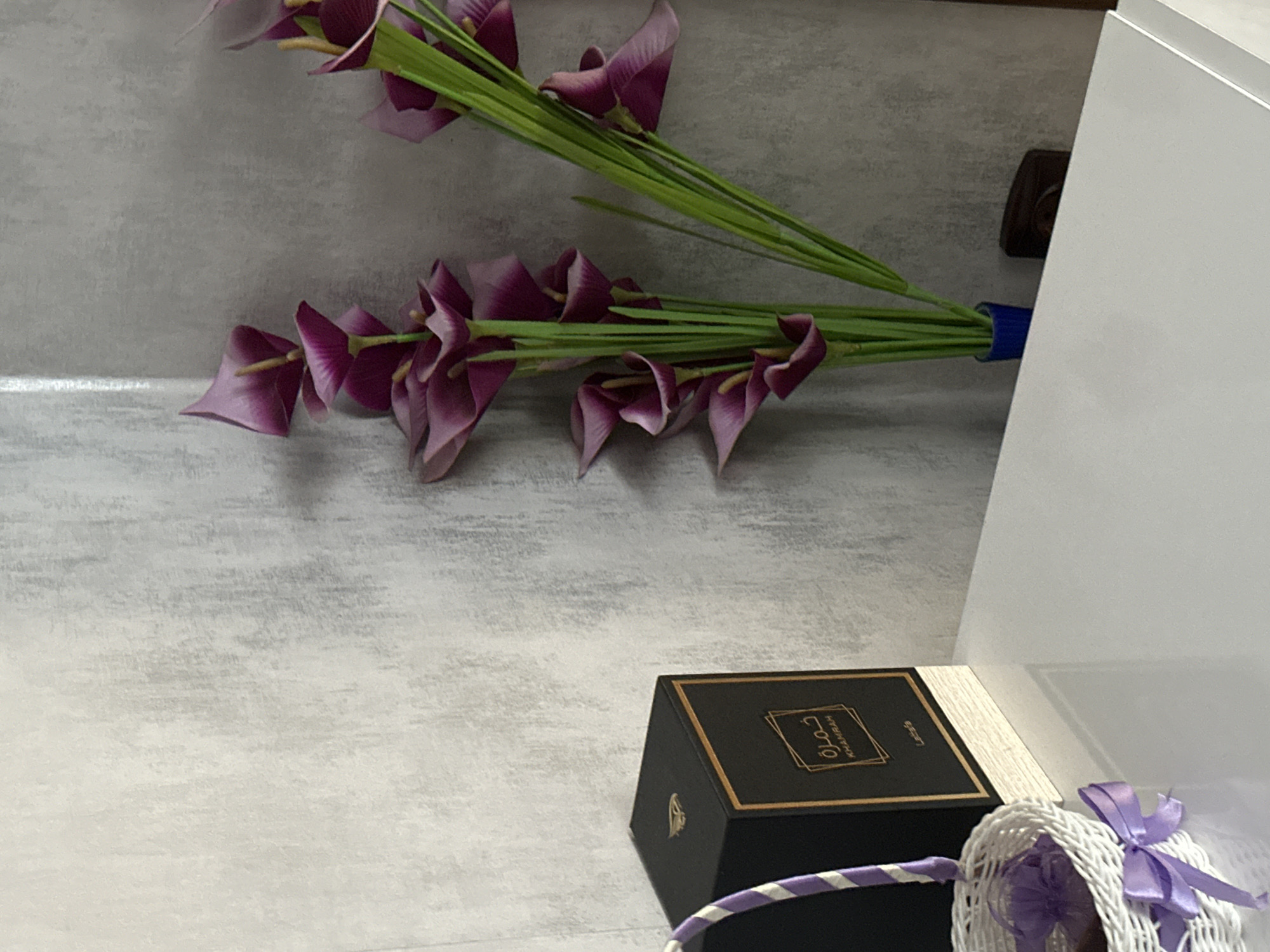 The image shows a still life arrangement on a light gray, textured surface that appears to be a shelf or tabletop. The scene includes artificial purple calla lilies, a black and gold box with Arabic script, and a small white wicker basket with a purple ribbon. The arrangement is placed adjacent to a white vertical surface, possibly a cabinet or wall. The location is not identifiable as Lovech, Bulgaria based on the given image.