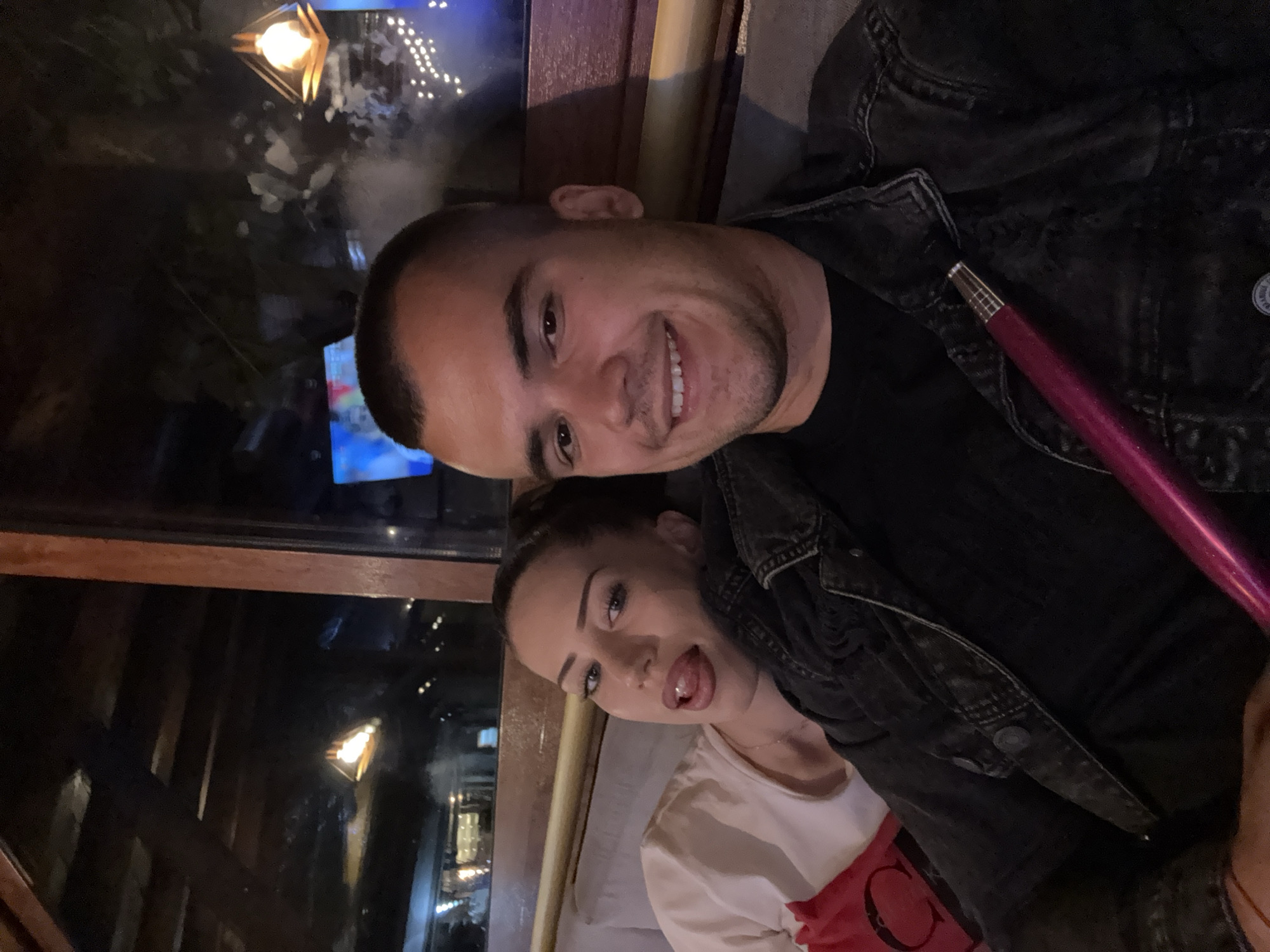 The image features a close-up, angled selfie of a young couple. The man, on the left, has short dark hair and is smiling broadly at the camera. He is wearing a dark denim jacket over a black t-shirt. The woman, on the right, is leaning in towards the camera with her tongue playfully sticking out. She has long dark hair and is wearing a light-colored t-shirt with a red graphic element visible.
They appear to be in an indoor setting, possibly a lounge or restaurant, indicated by the dark wooden interior and soft lighting. Reflections in what looks like a window or glass surface show glimpses of outdoor lights and possibly foliage, suggesting an evening or nighttime setting. A purple hookah hose is visible, held by the woman and extending towards the man's shoulder, hinting at the activity of smoking a hookah. The overall mood is casual and intimate, with both individuals expressing a sense of fun and connection. There is no visible text in the image.