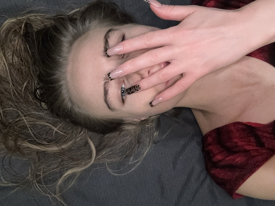 A woman lies on her back, partially covered by a gray blanket and wearing a red shirt. Her hair, a mix of blonde and brown, is styled upwards and to the side. Her right hand covers part of her face, drawing attention to her long nails, one of which is a leopard print design. Only one eye is visible, blue and staring directly at the camera.