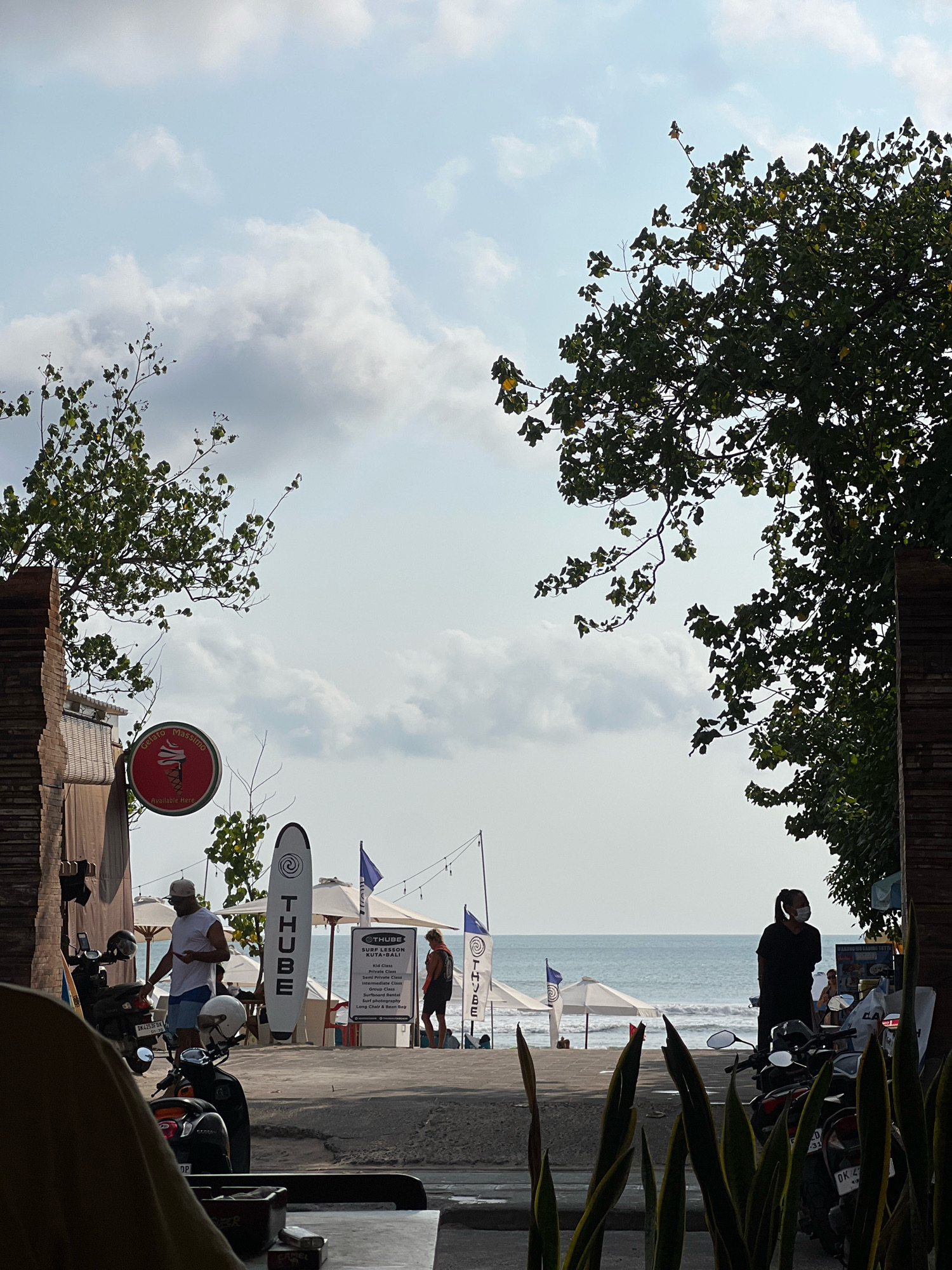 The image depicts a street scene in Kuta, Bali, Indonesia, overlooking the ocean. The foreground is dominated by the out-of-focus leaves of a snake plant, with a table and ashtray visible. In the mid-ground, several motorcycles are parked along a paved area. A man wearing a white t-shirt and a cap is standing near a motorcycle, holding a phone. Further back, a surfboard with the word "THUBE" is propped up. Beach umbrellas and flags are visible on the beach beyond the paved area. Two women are standing near the water's edge, one facing away from the camera and the other, wearing a mask, facing towards the camera. The ocean is visible in the background, with gentle waves. Trees with lush green foliage frame the top and right sides of the image. A red circular sign with an ice cream cone motif and the text "Gelato Massimo" is mounted on a brick wall on the left side of the frame. A sign for "THUBE SURF LESSON KUTA BALI" is also visible, listing various surf lesson options. The sky is partly cloudy with a bright, hazy quality.