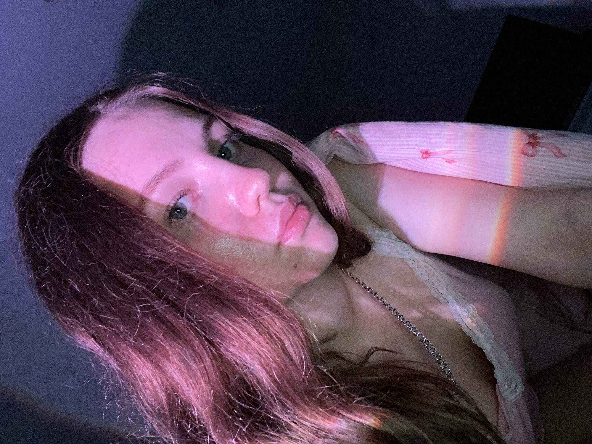 A young woman with long, dark hair illuminated by pink and purple light is the sole subject of this close-up photograph. She is positioned in the foreground, with her head tilted and her gaze directed slightly away from the camera. Her skin appears smooth, with a subtle sheen from the lighting. She is wearing a light pink top with white lace trim, and a silver chain necklace is visible. The background is dark and indistinct, suggesting an indoor setting. The lighting creates a dramatic effect, casting shadows and highlights across her face and hair. The image appears to be a selfie, taken at a slightly upward angle. There are no discernible elements in the image that indicate a specific location within the city of Izhevsk, Russia.