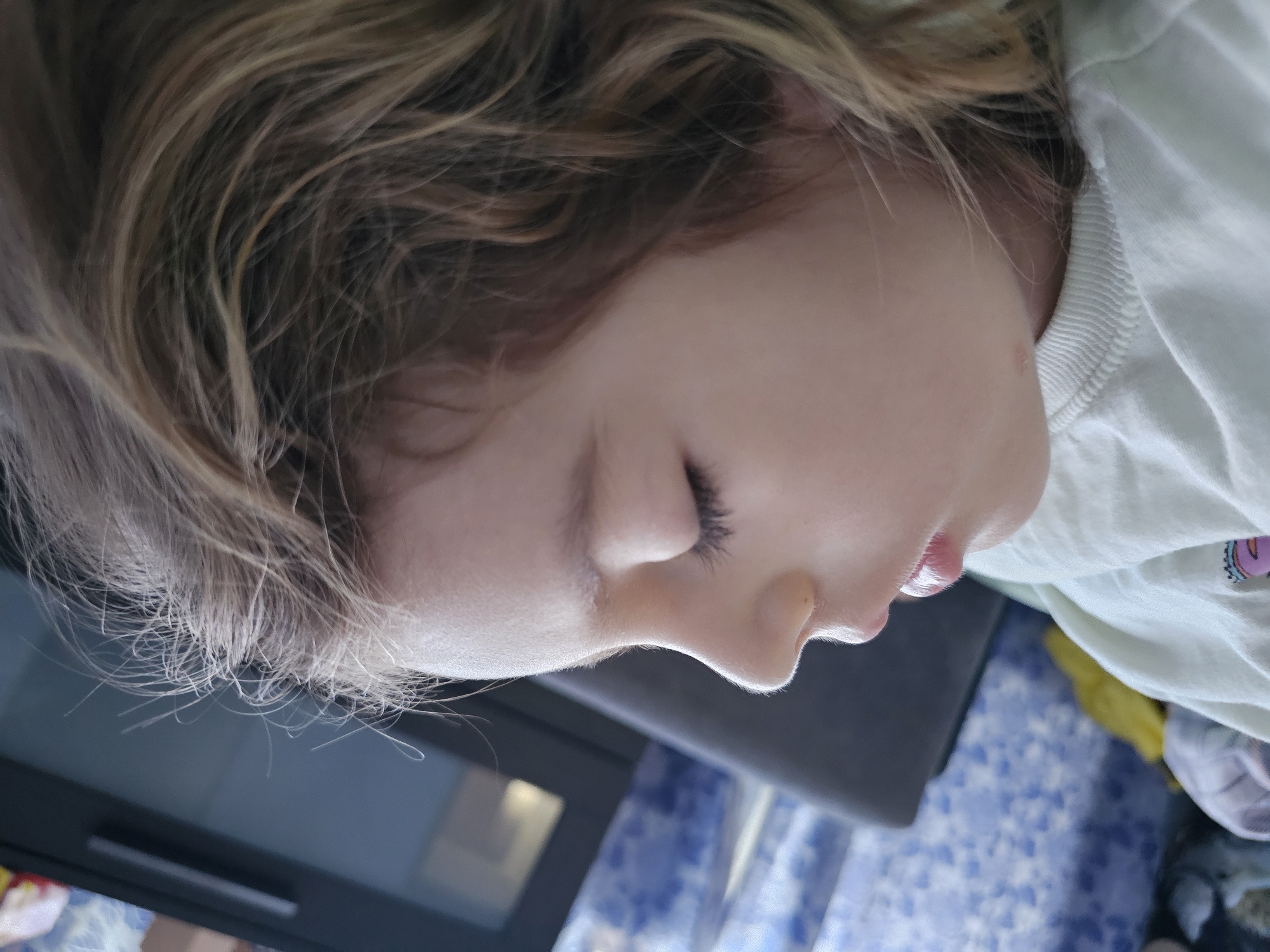 A close-up profile shot shows a young child with blonde hair and fair skin. The child's eyes are closed. The child is wearing a light-colored shirt. The background appears to be a room with blue patterned fabric and dark furniture. There is no indication this photo was taken in Khimki, Russia.