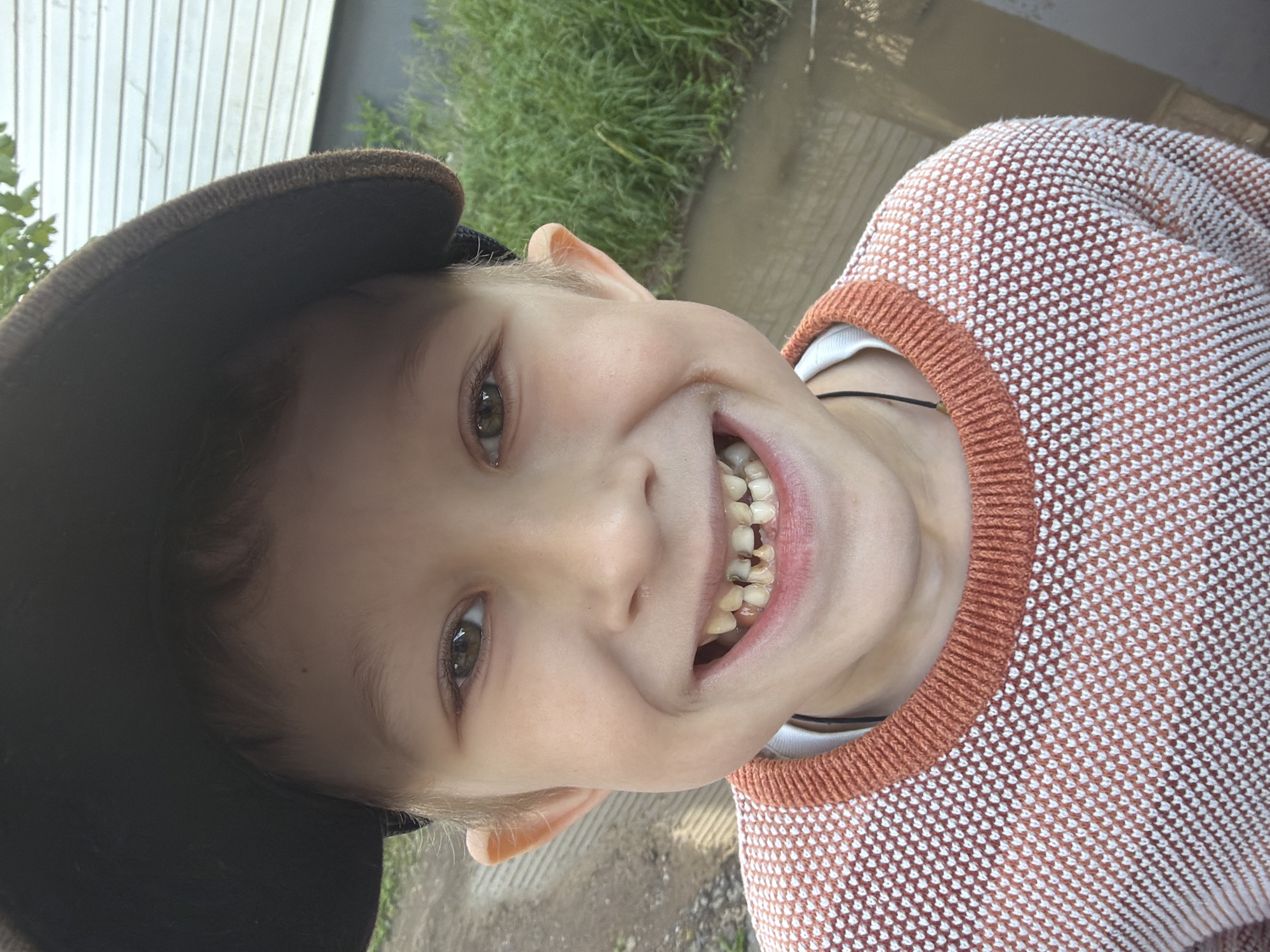 A close-up, rotated portrait captures a young boy smiling broadly and looking directly at the camera. He has fair skin and light-colored eyes, with some brown hair visible under a dark cap. He is wearing a white undershirt beneath an orange and white patterned knit sweater featuring an orange ribbed collar. His wide, joyful smile reveals his teeth, which show some discoloration or plaque.
The scene is set outdoors in Chaikovsky, Russia, illuminated by natural daylight. The background features green foliage or grass, along with what appears to be a light-colored building or fence. The ground beneath the boy seems to be dirt or an unpaved surface. The predominant emotion is one of happiness and joy, expressed through the child's bright eyes and wide smile. There is no visible text in the image.